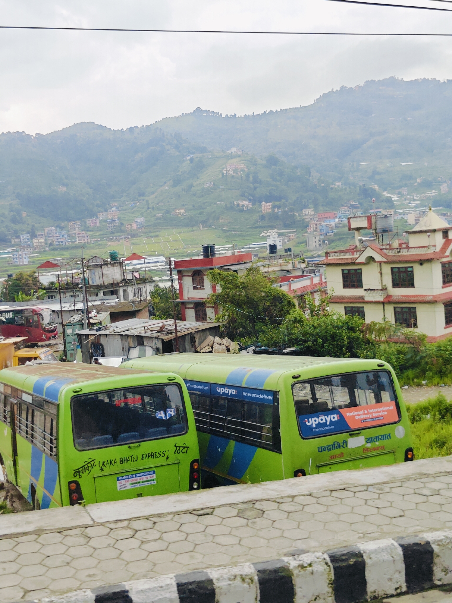 This is an eye-level, medium shot of two green and blue buses parked on a road in Chandragiri, Nepal. The buses are the primary focus of the image. The bus on the left is partially obscured by the bus on the right. Both buses have windows and signs on their rear. The bus on the right has "upaya #heretodeliver" and "National & International Delivery Service" written on it in blue and orange. Below that, in Nepali, it says "Chandragiri" and "Yatayat" (transportation). The bus on the left has "KAKA BHATIJ EXPRESS" written on its rear along with Nepali script. There are some electrical wires stretching across the top of the image. The background is a hazy hillside covered in houses and greenery, suggesting a mountainous urban or semi-urban environment. The overall impression is a typical scene of public transportation in a hilly Nepalese city.