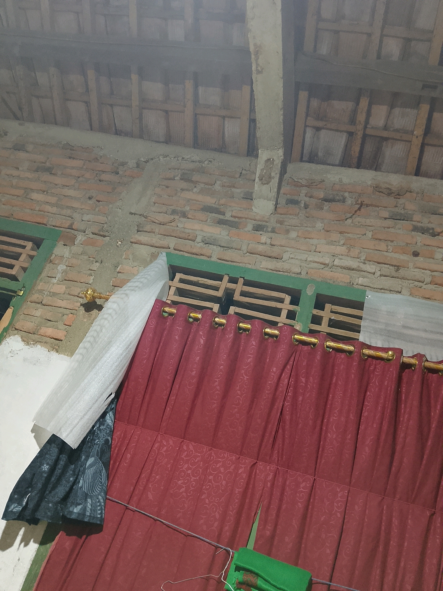 The image depicts an interior view of a building under construction or renovation in Negeri Agung, Indonesia. The foreground is dominated by red curtains with a subtle embossed pattern and gold grommets, hung from a rod. To the left of the red curtains, a partially visible dark-colored patterned fabric is draped, and a white, textured material is also present. A thin wire runs horizontally across the lower portion of the image, with a green folded material and a metal hanger visible near it. Above the curtains, a brick wall is visible, constructed with uneven reddish-brown bricks and some sections of grey concrete mortar. Further up, a wooden ceiling structure with visible beams and planks is present. The overall impression is that of an unfinished or functional space.