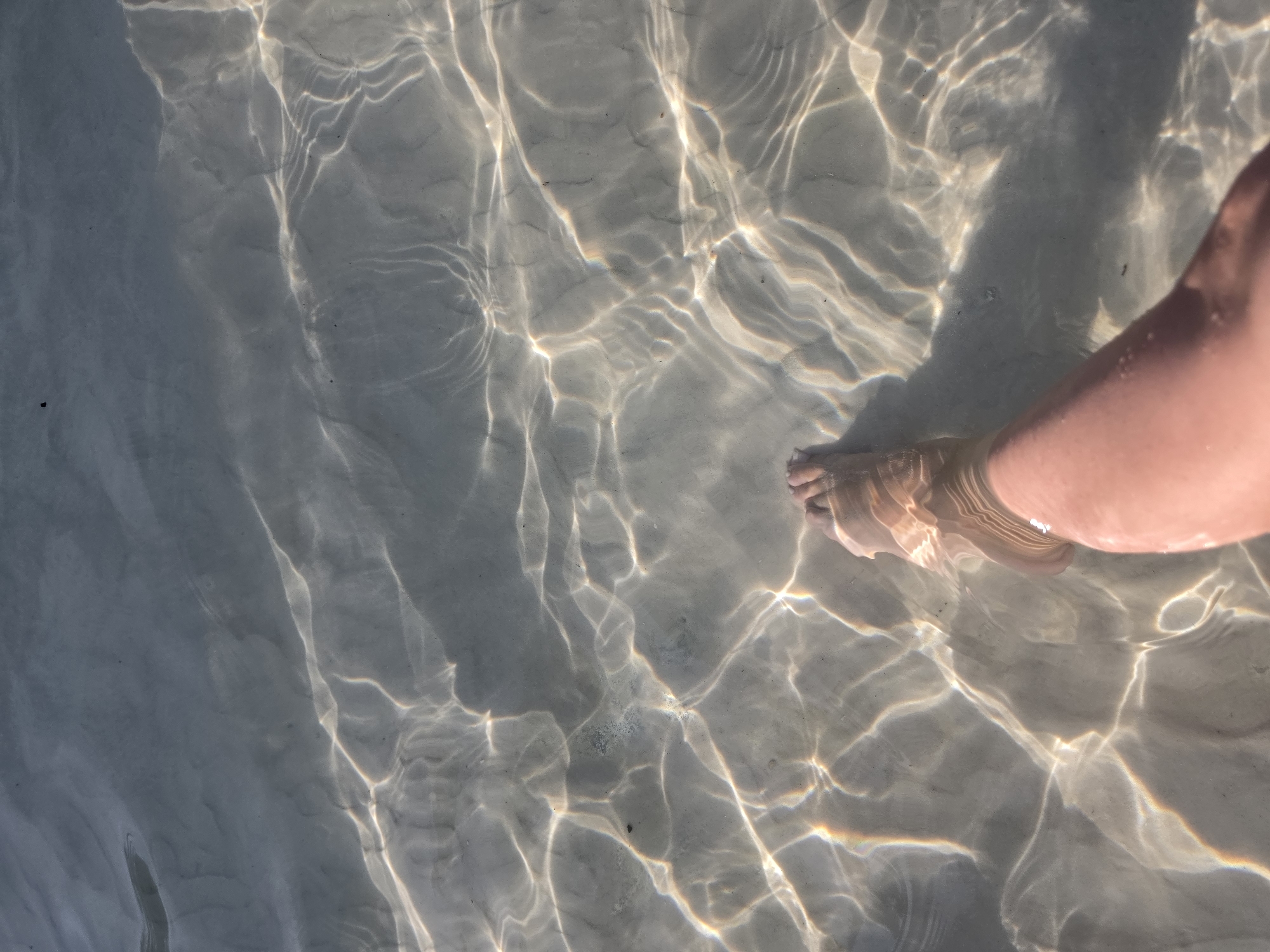 The image shows a person's leg and foot submerged in shallow, clear water. The water is transparent, revealing a sandy bottom with patterns of light and shadow created by the surface ripples. The light refracts and reflects off the water, creating bright, shimmering lines and shapes on the sandy seabed. The leg and foot appear to be a pale skin tone, and the toes are visible through the water. The overall scene suggests a serene and natural environment, possibly a beach or shallow body of water. Given the prompt's request for Jesolo, Italy, this scene could depict a shallow area along the coast of Jesolo.
