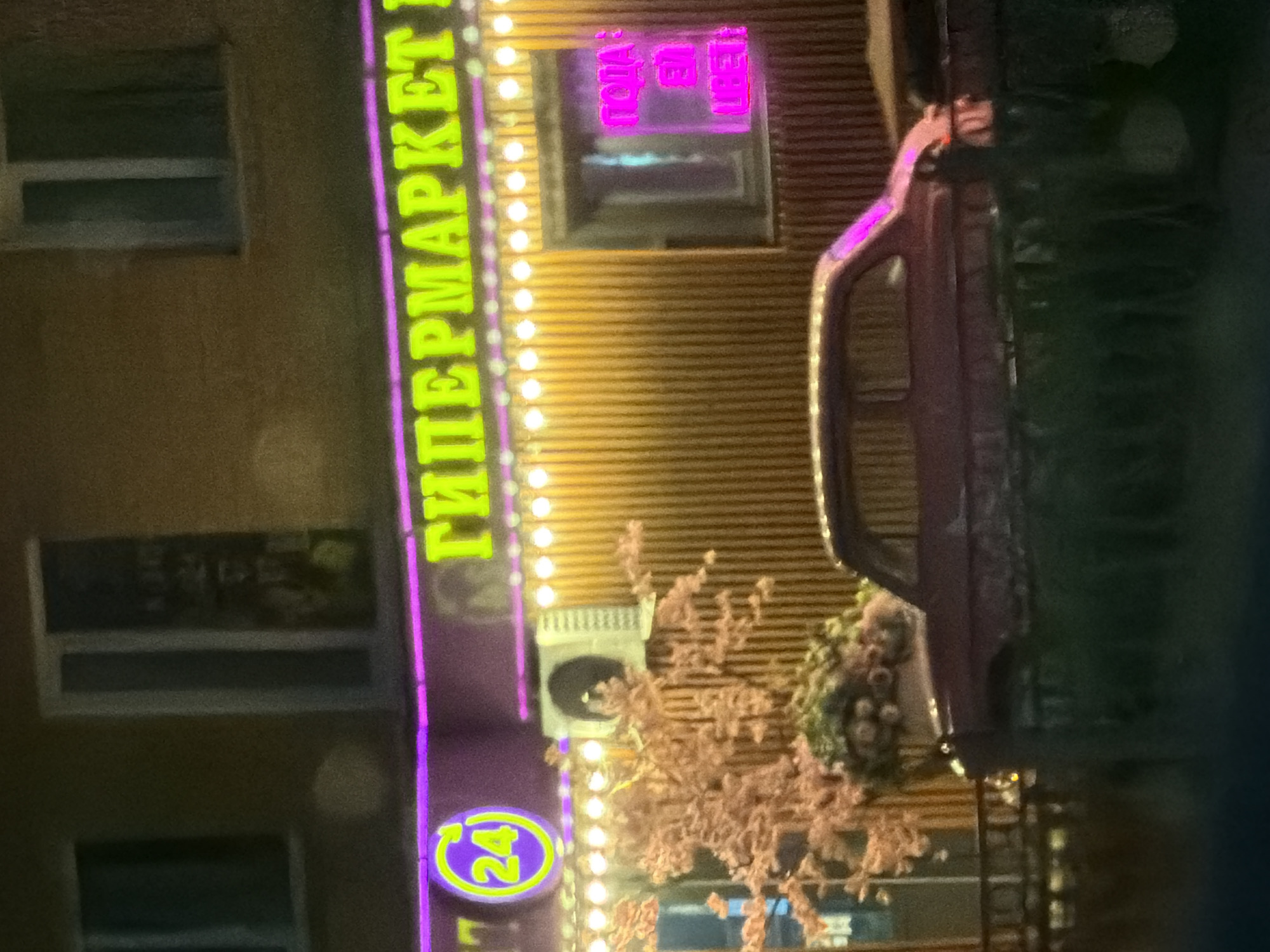 The image displays the facade of a "Гипермаркет" (Hypermarket) in Yekaterinburg, Russia, illuminated by neon lights. A prominent sign in neon green letters reads "ГИПЕРМАРКЕТ", with a circular "24" logo in purple and green on the left. The building has several windows and is adorned with a string of small, bright lights. An air conditioning unit is mounted on the wall. In front of the building, a vintage red car is parked, and decorative pink flowering branches are visible. The overall impression is of a commercial establishment at night, likely in a somewhat older part of the city given the car's style.