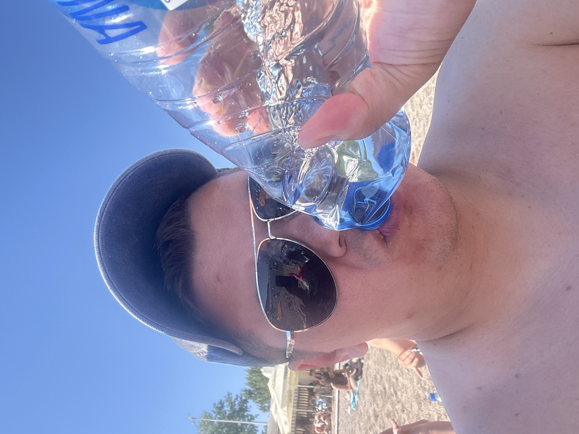 A man, wearing sunglasses and a cap, drinks water from a clear plastic bottle. He is shirtless and on a beach, as evidenced by the sand and people in the background. The scene appears to be outdoors under a clear blue sky. The background shows a beach with umbrellas and people lounging. The location is the city of Prijevor, Montenegro.