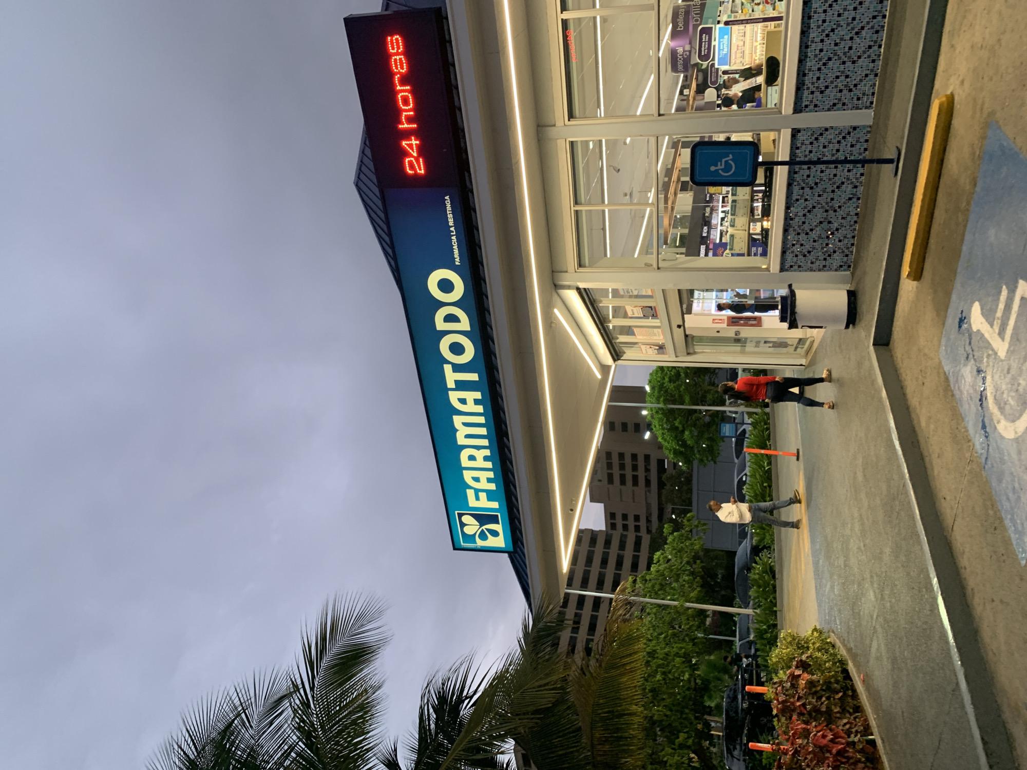 The image captures the exterior of a modern "Farmatodo" pharmacy in Pampatar, Venezuela, at dusk or early evening under an overcast sky. The building is brightly lit, contrasting with the diminishing daylight.
A prominent vertical sign on the building features illuminated text. At the top, "24 horas" is displayed in red, indicating its operating hours. Below this, "FARMACIA LA RESTINGA" is visible in smaller text. The main part of the sign showcases the large "FARMATODO" brand name in a blue-green color with its distinctive logo.
The building has large glass windows, revealing the store's interior with shelves stocked with products, some labeled "belleza" (beauty), "personal," and "familia." Near the entrance, a blue sign with the international symbol of access (handicapped) is displayed.
On the concrete walkway outside the store, two individuals are visible. One person, dressed in a red top and dark pants, walks away from the entrance, while another, wearing a white shirt and dark pants, is closer to the building. The ground features a blue painted handicapped parking space symbol and a yellow concrete parking curb.
The scene is framed by tropical vegetation, including several large palm trees in the foreground and surrounding the building. In the background, other multi-story buildings are visible, suggesting an urban or semi-urban setting.