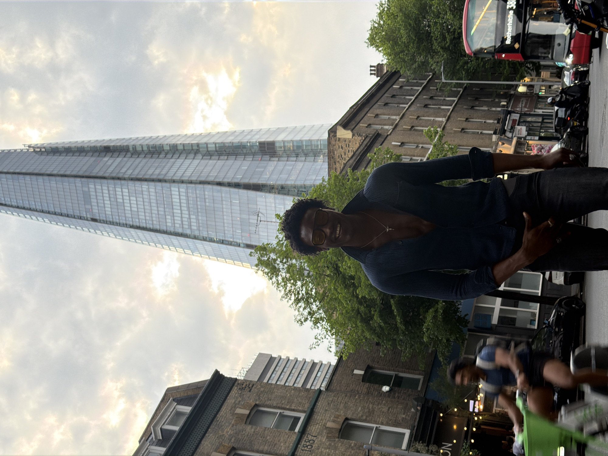The image displays a lively urban scene in London, United Kingdom, featuring a person in the foreground set against a backdrop of prominent city architecture and street activity.
In the foreground, a person with dark, curly hair and glasses is smiling and looking directly at the viewer. They are wearing a dark blue long-sleeved shirt and dark trousers, with a slender necklace visible.
Behind the person, the striking, modern glass skyscraper known as The Shard dominates the left side of the background, its reflective surface blending with the sky. To the right of The Shard, a series of older, multi-story brick buildings with numerous windows line the street. One of these traditional buildings, closer to the foreground, has "1887" inscribed on its facade.
Further into the background on the right, a red double-decker bus, characteristic of London, is partially visible, with its destination board clearly displaying "Rotherhithe 381." Several motorcycles or scooters are parked along the street in front of the brick buildings. A blurry figure on a bicycle or scooter is also visible in the lower right, suggesting motion. Green trees are interspersed among the buildings, adding natural elements to the urban environment.
The sky is mostly cloudy with bright, diffused light, indicating a soft illumination, possibly during late afternoon or a bright overcast day. The overall scene captures the dynamic blend of historic and contemporary elements in a bustling city setting.