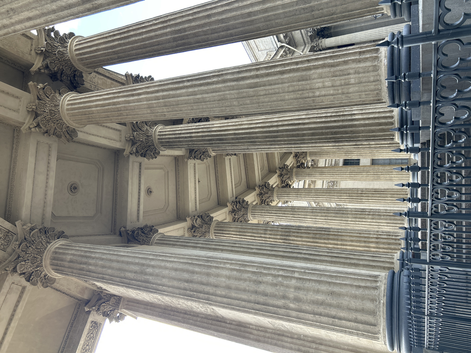 This is a low-angle shot of architectural details of a building in Saint Petersburg, Russia. The scene focuses on a series of tall, fluted columns that lead the eye upwards. Each column has an ornate capital, adorned with decorative foliage. The ceiling above the columns is decorated with recessed panels and intricate trim. A wrought-iron fence with pointed finials is seen in the foreground, on the right. The image is slightly overexposed, creating a bright and airy feel. The sky is visible in the gaps between the columns.