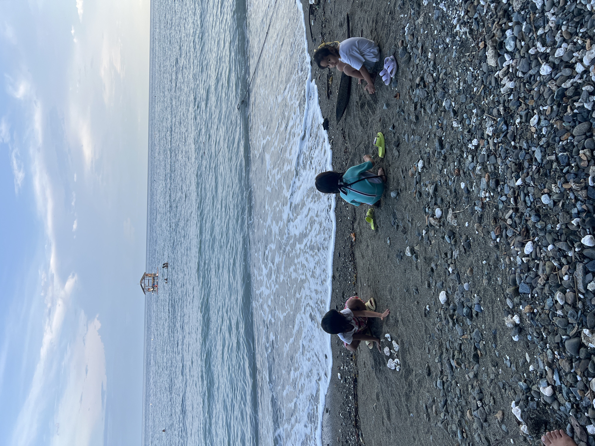 Three children are playing on a beach in Banaybanay, Philippines. The beach is a mix of sand and pebbles. The water is coming to shore with white foam. In the distance, there is a structure on the water. The sky is light blue. One child is wearing a white shirt and sandals, another is wearing a teal swimsuit and green sandals, and the third child has a white and floral-patterned shirt.