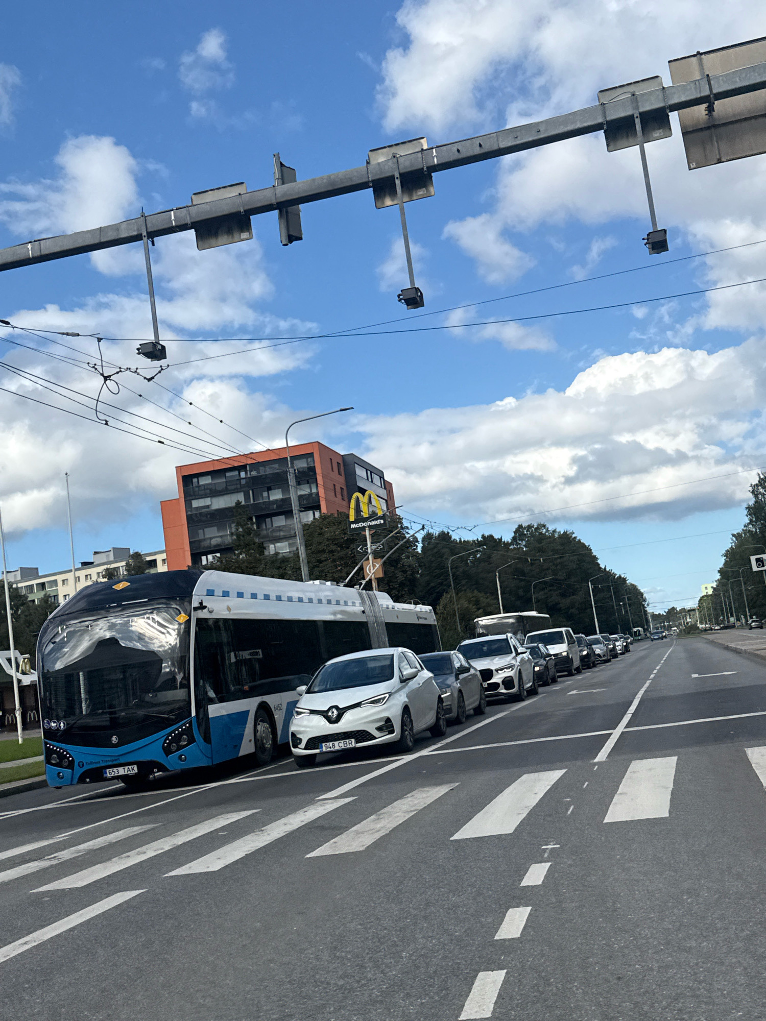 The image shows a traffic jam on a street in Tallinn, Estonia, under a partly cloudy sky. A modern blue and white trolleybus is in the foreground, stopped in traffic. Several cars are lined up behind it. To the left of the road, there is a tall, orange and black apartment building with many windows. A McDonald's restaurant sign is visible above the building. Overhead, traffic light poles and overhead wires for the trolleybus are present. The road has crosswalks and lane markings. The overall scene depicts a typical urban street with moderate traffic.