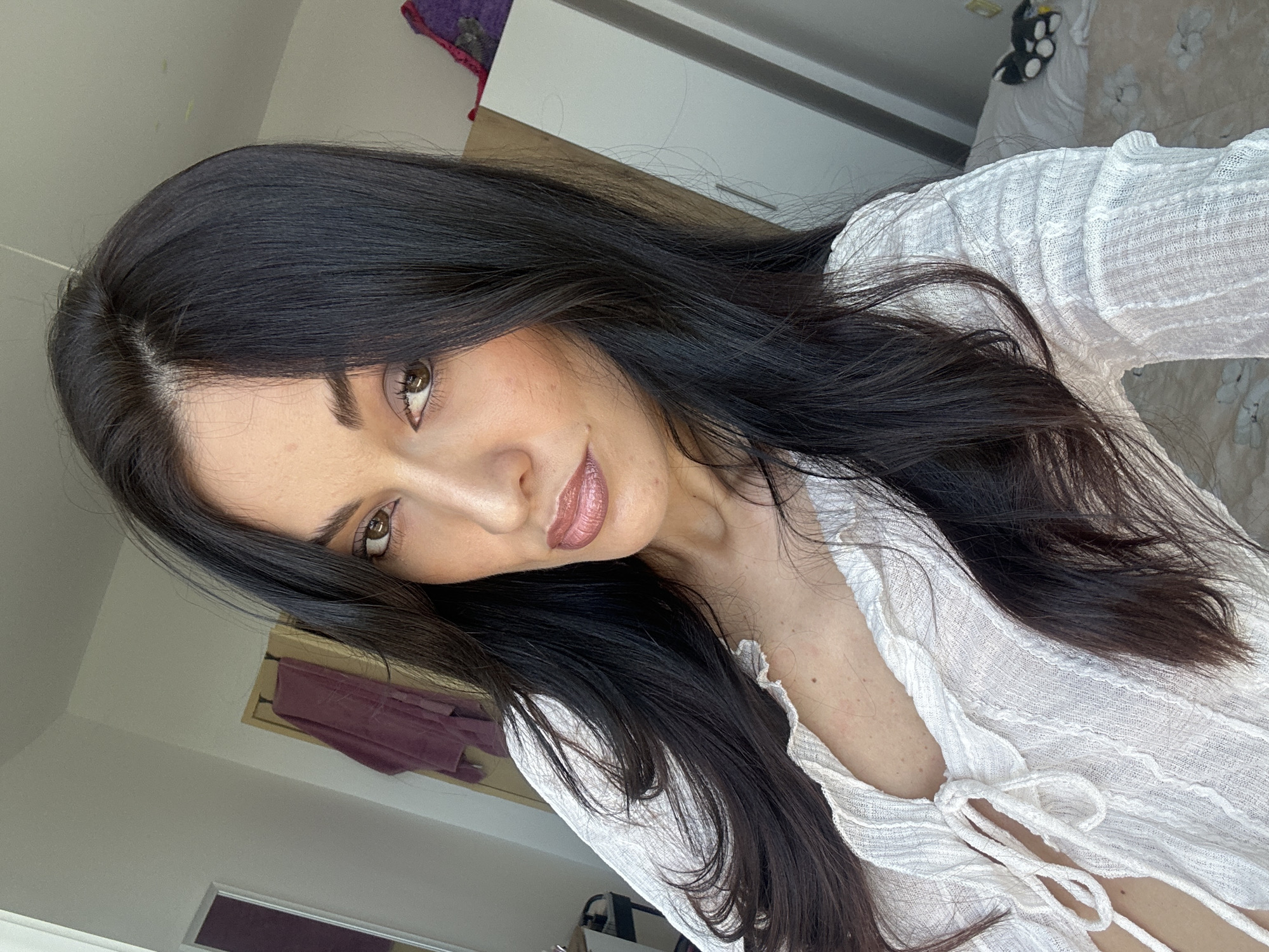 This is a selfie taken indoors, likely in a bedroom. The subject is a woman with long, dark hair and brown eyes, wearing a white, open-neck top. She is looking directly at the camera with a neutral expression. The background includes a wardrobe, a door with a draped garment, and a bed with bedding. There is no indication of the city of Burgas, Bulgaria in the image.