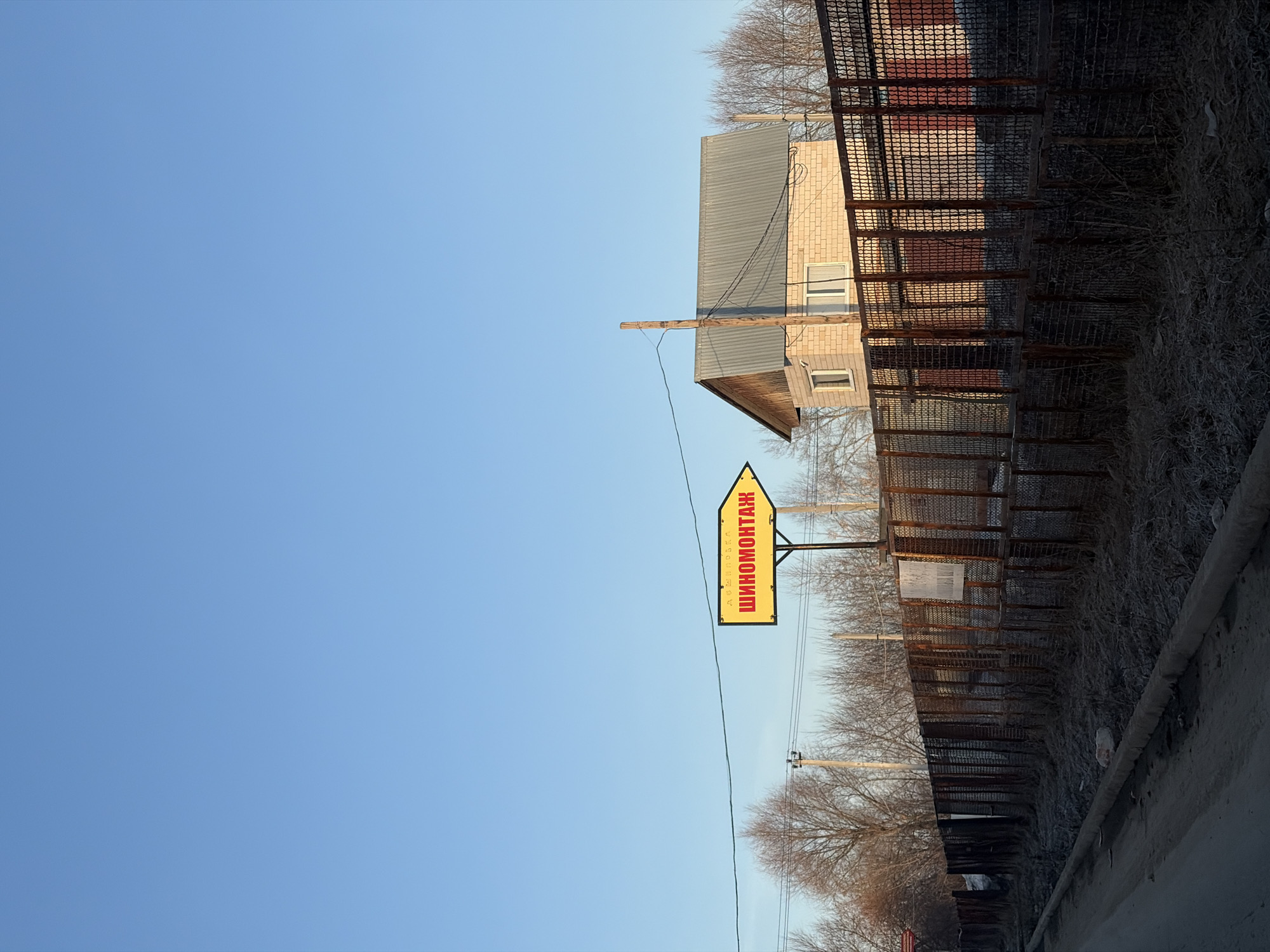 The image presents an outdoor scene, likely an urban periphery in Chaikovsky, Russia. In the foreground, there is a road or paved area with a partially visible curb. A rusty, wire-mesh fence runs along the right side, bordering a somewhat barren area with dry grass and weeds. Beyond the fence, deciduous trees are visible.
A yellow sign bearing text in Cyrillic, "ШИНОМОНТАЖ" (SHINOMONTAZH) is mounted on a pole. A utility pole stands to the right of the sign, with power lines stretching across the frame.
In the background, a light-colored brick building with a metal roof can be seen. The sky above is a clear blue.
The scene suggests an area where automotive repair services (tire fitting/repair - "ШИНОМОНТАЖ") are located on the outskirts of the town. The general impression is one of a somewhat run-down, post-industrial urban landscape.