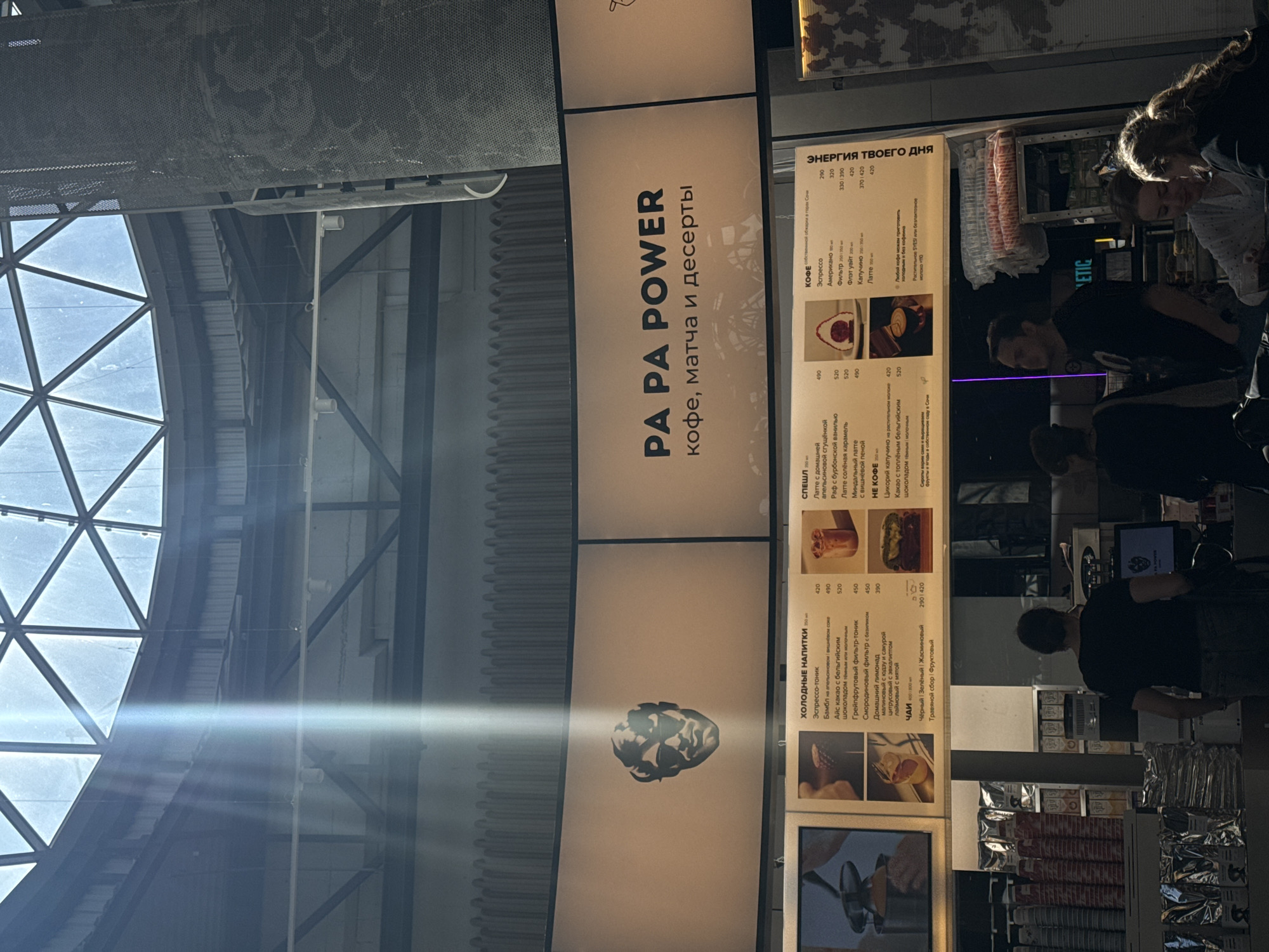 This is a vertical, eye-level shot of people interacting in what appears to be a modern, indoor space, possibly a cafe or retail establishment. In the foreground, three individuals are visible. The person on the far right, a woman with dark hair, is leaning over and looking down, possibly at a menu or item. Next to her, another woman, also with dark hair, is also looking down. A man in a black t-shirt stands in the mid-ground, his back to the camera, appearing to work or interact with a counter.
A large, illuminated menu board dominates the center of the image. The text on the board is in Russian and lists various beverages and desserts. Prominent headings include "PA PA POWER кофе, матча и десерты" (PA PA POWER coffee, matcha, and desserts) and "ЭНЕРГИЯ ТВОЕГО ДНЯ" (Energy of your day). Other sections detail "КОФЕ" (Coffee), "СПЕШЛ" (Special), "ХОЛОДНЫЕ НАПИТКИ" (Cold Drinks), and "ЧАИ" (Tea), along with prices. Images of food items are also displayed on the menu.
The background suggests a spacious interior with industrial elements, including a metal grid ceiling and exposed beams. Large windows or a skylight, forming a triangular geometric pattern, are visible in the upper left, letting in a bright, possibly daytime light, which creates a lens flare effect across the image. The overall atmosphere appears to be casual and social.