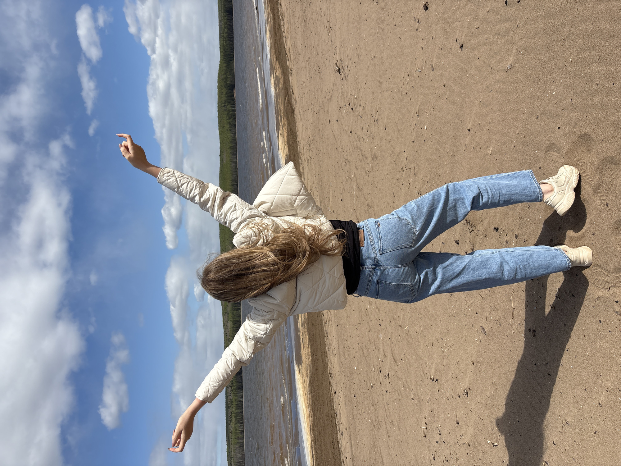 A young woman lies on her back on a sandy beach, arms outstretched to the sides. She wears a light-colored quilted jacket, a black top, light blue jeans, and white sneakers. Her blonde hair is spread out behind her. The background consists of water, a tree line, and a blue sky with white clouds. The location is a beach in the city of Chaikovsky, Russia.