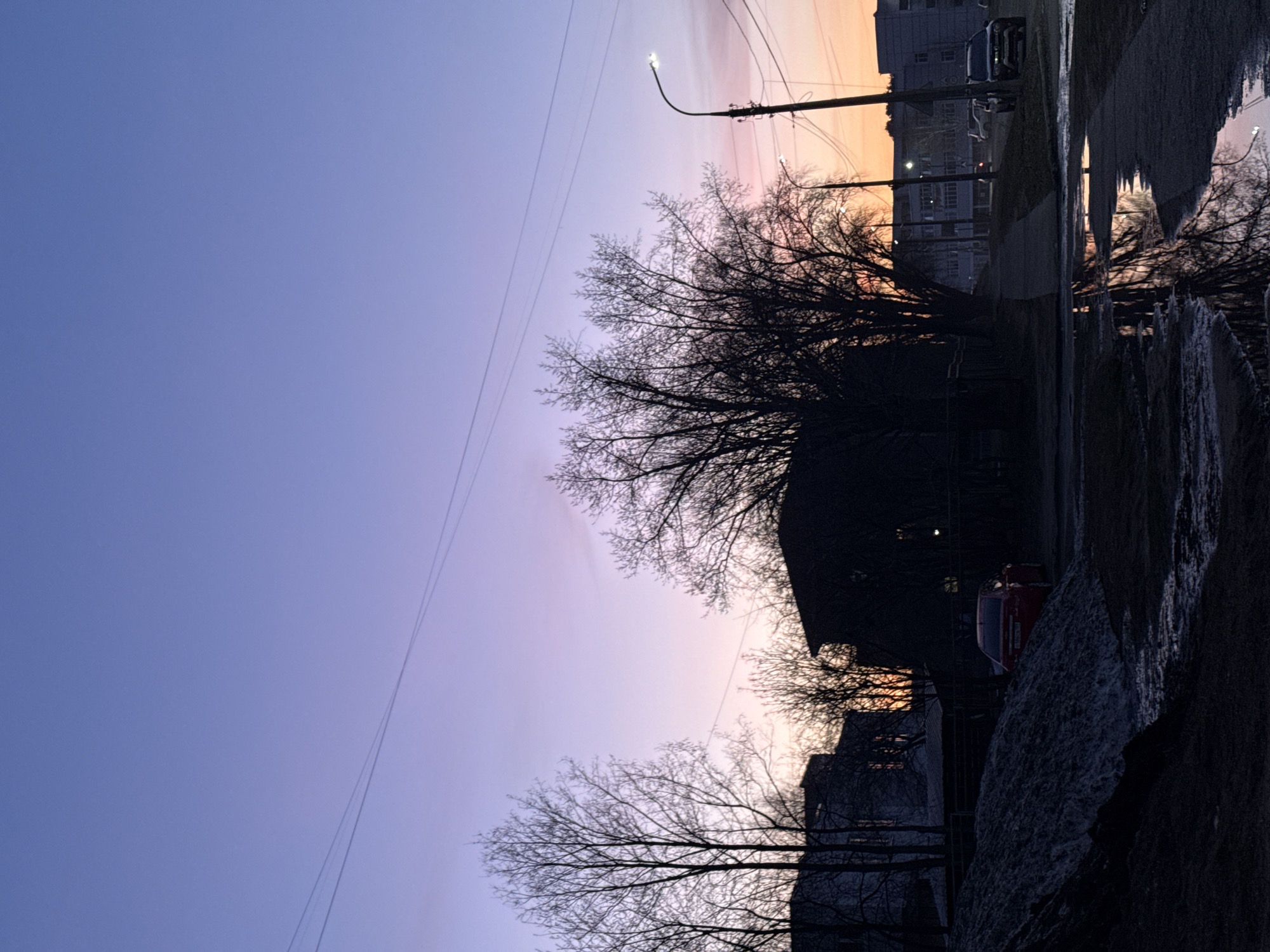 The image depicts an outdoor, urban or suburban scene captured at either dawn or dusk, as indicated by the vibrant, multi-hued sky and artificial lights. The sky transitions from a deep, clear purple-blue at the top-left to softer lavender and then to warm orange and pink hues towards the horizon on the right.
Silhouetted against this colorful sky are numerous bare trees, their intricate branches reaching upwards, suggesting either winter or early spring. Several utility poles and a network of overhead wires crisscross the frame. Illuminated streetlights are visible, one prominently curving over the street on the right.
The lower half of the image features the ground, which appears wet and reflective, possibly from recent rain or melting snow. These wet surfaces mirror the sky's colors and the glow of the streetlights. Dark, shadowed buildings, some with visible rooflines and subtle architectural details, line the right side of the street. Two vehicles are visible: a dark SUV or van parked on the far right, and a red car partially visible further down the street to the left. The red car has a license plate with unreadable Cyrillic characters.
The scene conveys a quiet, tranquil atmosphere typical of the transitional moments between day and night in a residential or mixed-use area in Chaikovsky, Russia. There are no people visible, and no specific activity is taking place, contributing to the peaceful and still quality of the image.