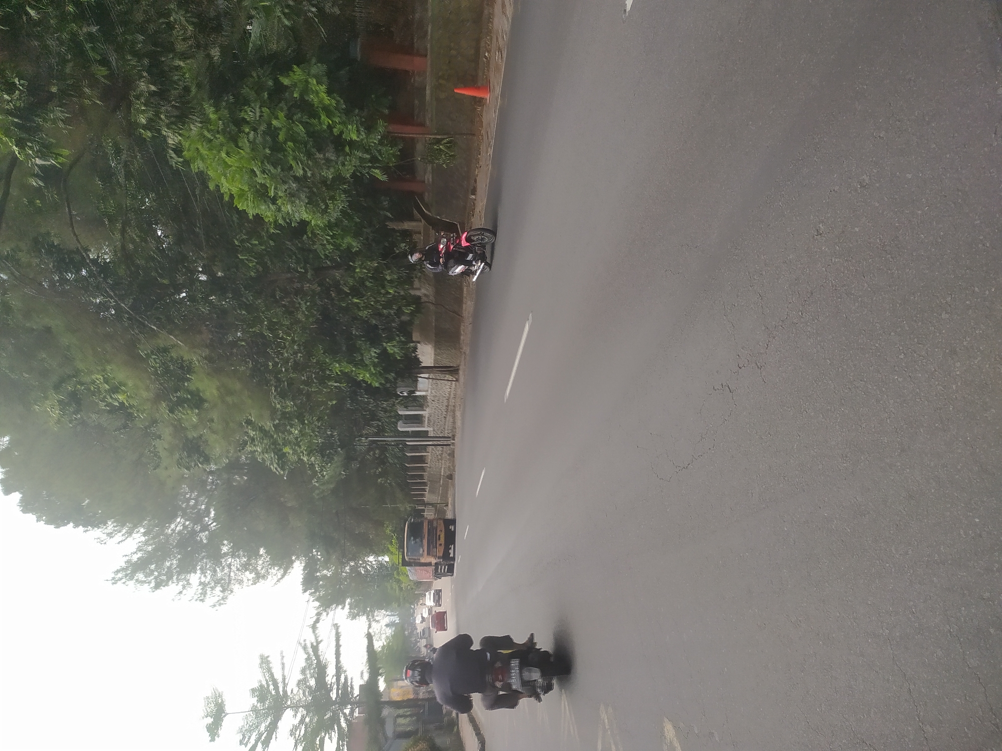 The image captures a street scene in Salatiga, Indonesia, on a hazy day. In the foreground, a man on a black motorcycle is riding away from the camera, wearing a dark shirt and a helmet. The motorcycle has a license plate that reads "H 3258 AP". Further down the road, a pink motorcycle with a rider wearing a helmet and a dark jacket is also moving in the same direction. On the right side of the road, an orange traffic cone is visible. In the background, a large orange truck is driving on the road, and several cars are also present. The street is lined with lush green trees and a stone wall. The sky is overcast, creating a diffused light.