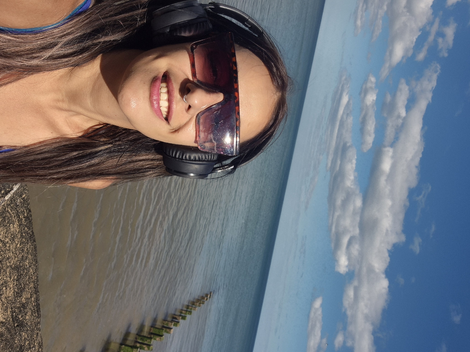 A woman with long, dark hair, wearing large headphones and tortoiseshell sunglasses, takes a selfie by the sea. She has a nose ring and is smiling. Behind her, the calm sea stretches to the horizon under a bright blue sky dotted with white clouds. A line of wooden posts, covered in green algae, protrudes from the water in the distance. The location is Bridlington, United Kingdom.