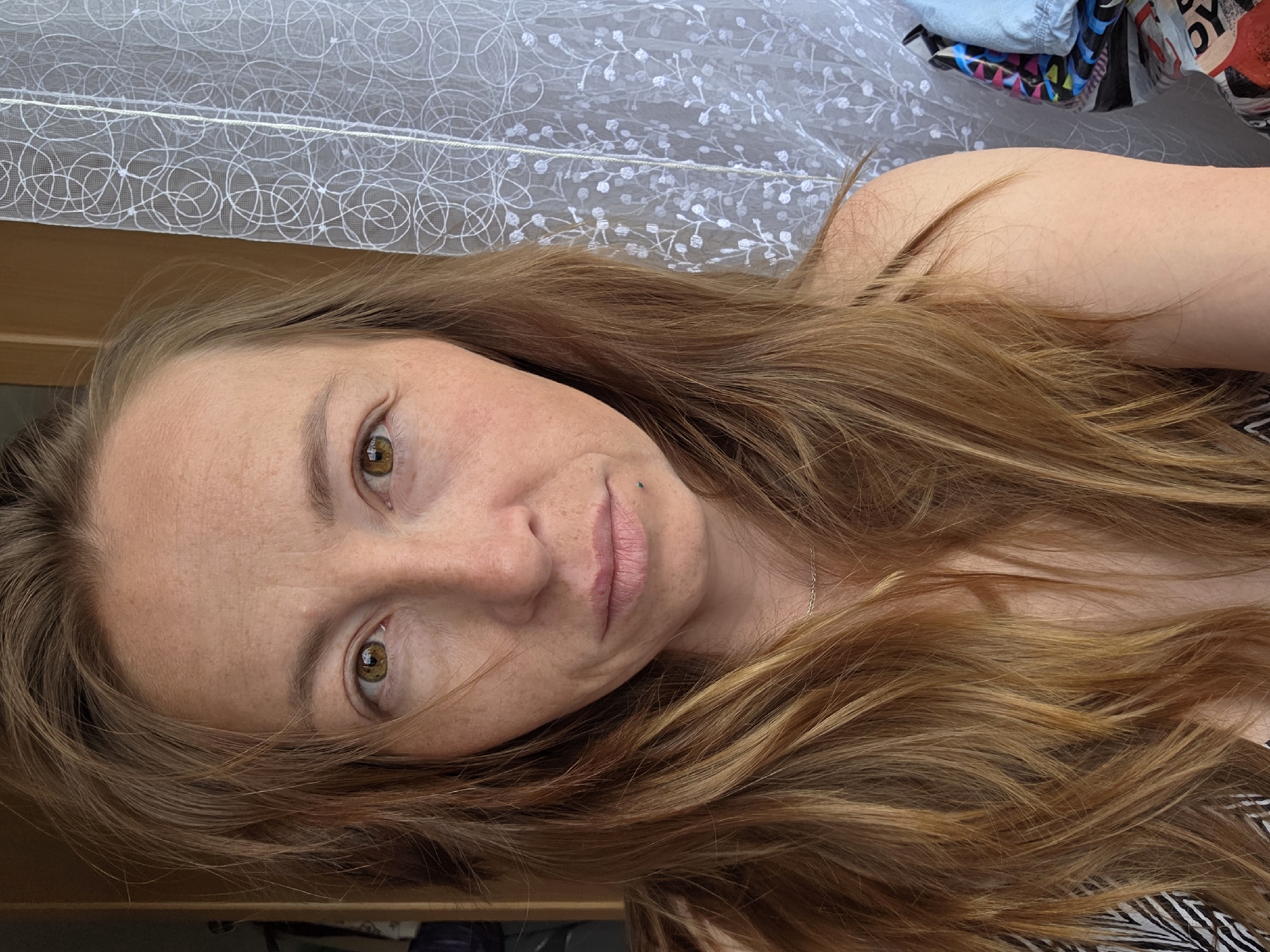 A head-and-shoulders portrait captures a woman lying down indoors, looking directly at the camera with a neutral expression. She has long, wavy reddish-brown hair, visible around her face and draped over her shoulder. Her skin is light-toned with visible freckles, particularly across her nose and cheeks. Her eyes are a striking green-brown or hazel. A small, dark piercing or mole is visible near the left corner of her mouth (viewer's right). A thin, silver-colored necklace chain is barely visible against her skin.
The background primarily consists of a white, sheer curtain with a swirling, circular pattern on the left, transitioning to a floral or leafy pattern on the right. Below the curtain, a light-colored wooden surface is partially visible. Further in the background, behind her right shoulder, there's a glimpse of what appears to be rumpled fabric or clothing, including a light blue garment and other colorful patterned items. The setting appears to be a casual indoor space, possibly a bedroom, indicated by the curtain and relaxed pose. The lighting suggests daytime, with natural light coming from behind or above her. No discernible text is visible in the image. The overall mood is calm and personal.