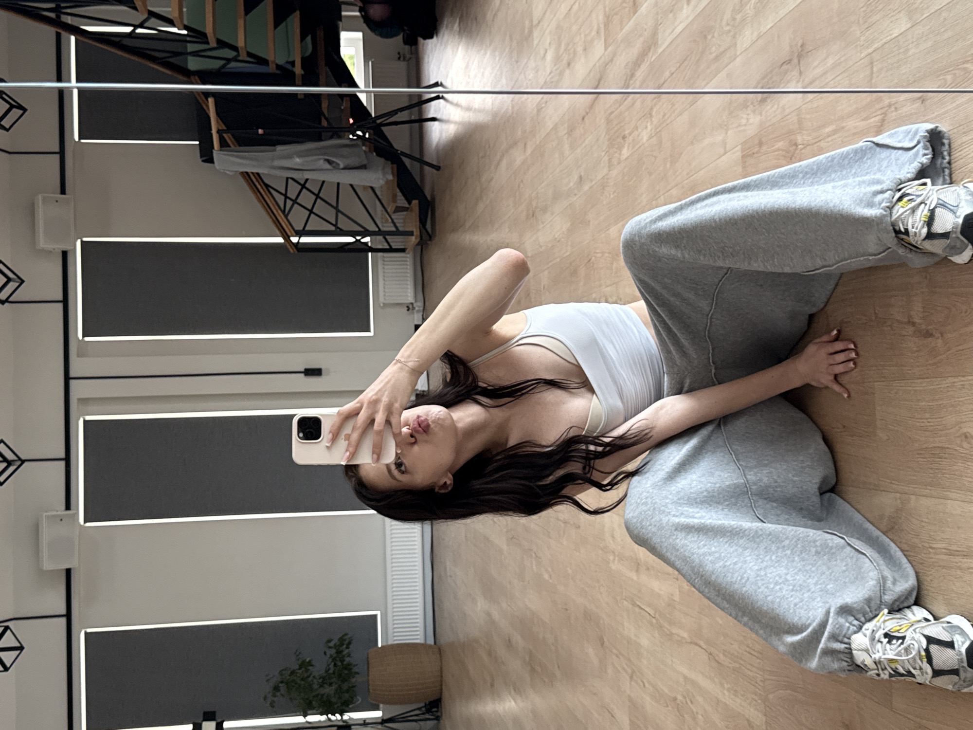 A young woman with long, dark hair is sitting on a wooden floor in what appears to be a dance studio or a gym. She is taking a selfie with her phone, which she is holding up to her face. She is wearing a white tank top and loose-fitting gray sweatpants. Her legs are spread wide apart, and she is looking at the phone with her lips puckered. In the background, there are large windows with blinds, a plant in a basket, and a staircase. The lighting is bright, and the overall scene suggests a casual and relaxed atmosphere. The location is Agalarov Estate, Russia.