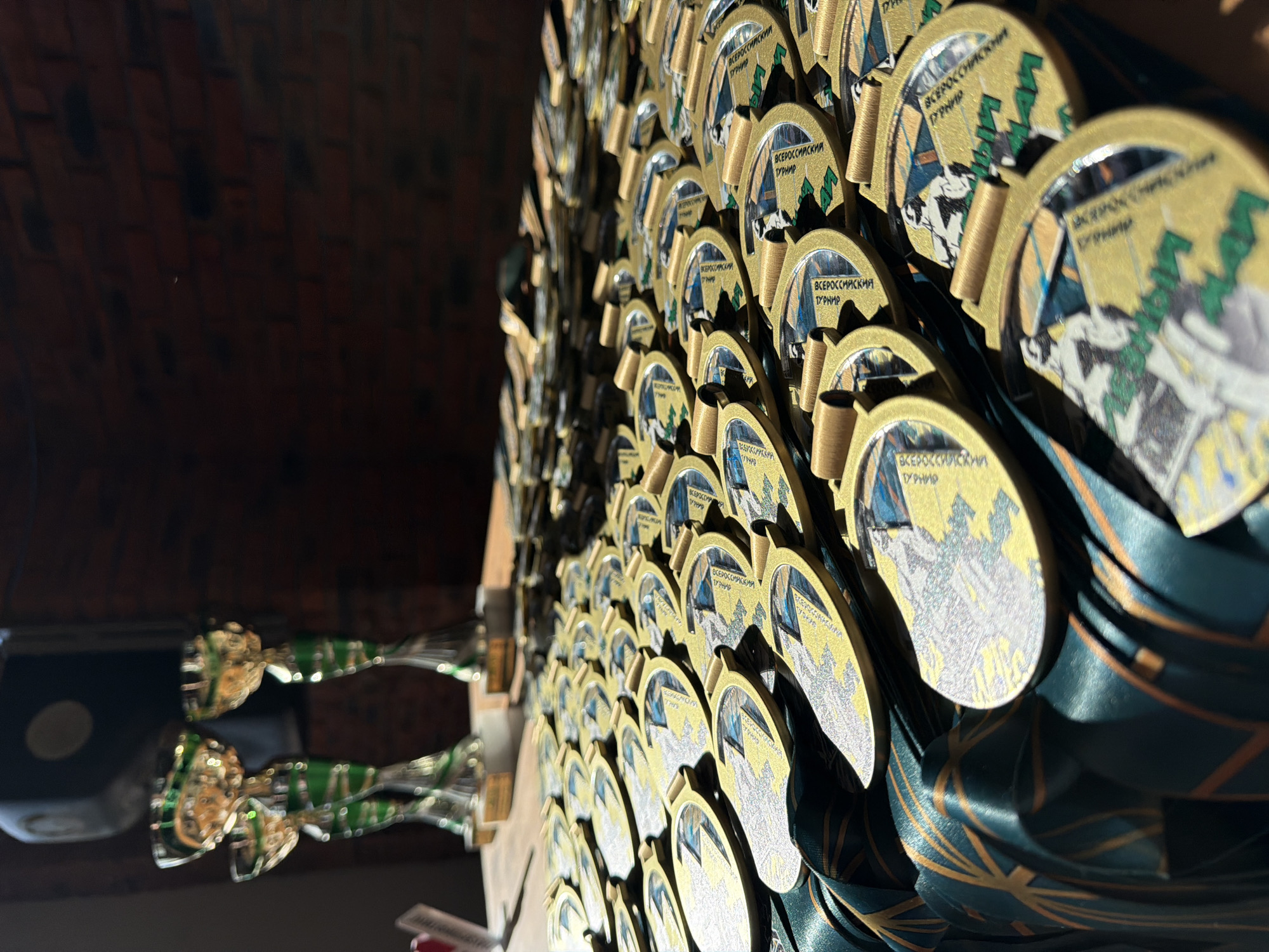 The image shows a close-up, slightly tilted view of a display of medals and trophies, seemingly indoors. The medals are round, predominantly gold in color, and feature a design including an illustration of a snowy mountain scene and a circular text element in Russian - "ВСЕРОССИЙСКИЙ ТУРНИР" meaning "All-Russian Tournament". The medals are attached to dark green ribbons with gold geometric patterns.
To the left of the frame, slightly out of focus, are two trophies with gold and green accents, sitting on a dark surface. The background consists of a dark brick wall, indicating an indoor setting, possibly in the city of Chaikovsky, Russia.