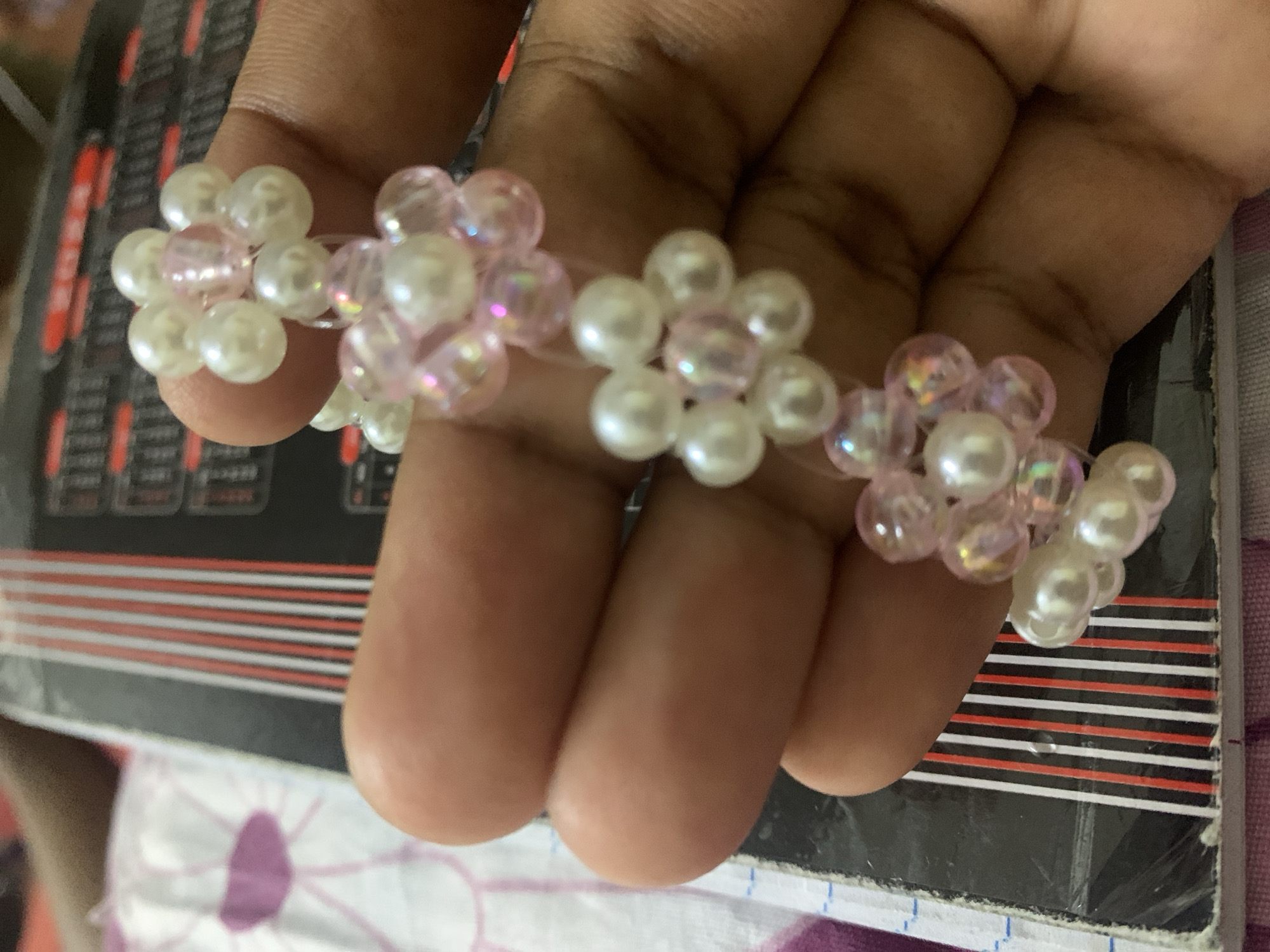 A close-up shot shows a person's hand holding a beaded bracelet made of faux pearl beads and pink iridescent beads. The beads are arranged in a floral pattern. The bracelet is placed across the fingers. The background includes a black object with red and white stripes, and a white surface with purple floral designs. The location is not discernible from the image.