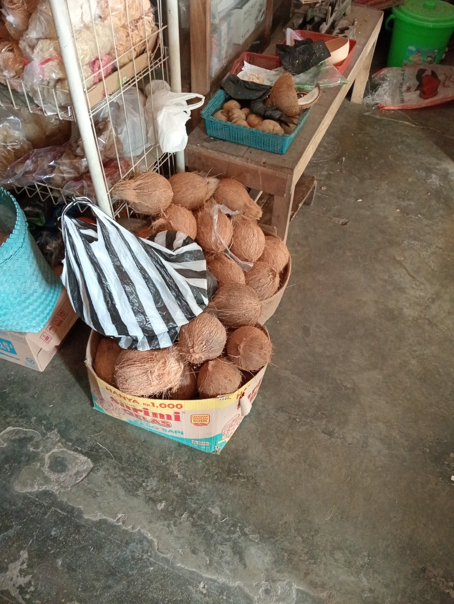The image depicts a retail scene likely in Grabag, Indonesia, featuring a variety of goods displayed in a somewhat disorganized manner. In the foreground, a large cardboard box is filled with coconuts. A black and white striped bag is placed on top of the coconuts within the box, obscuring some of them. To the left of the box, a portion of a light blue woven basket is visible. In the background, a metal shelf unit holds various bagged food items. A wooden table with several items on it, including a blue plastic basket containing small round objects and what appears to be ginger or garlic, is positioned to the right of the shelf. A green plastic bucket is also visible in the upper right corner. The floor appears to be concrete, with some scuff marks and stains. The lighting suggests an indoor setting, possibly a market stall or small shop. No people are visible in the image.
