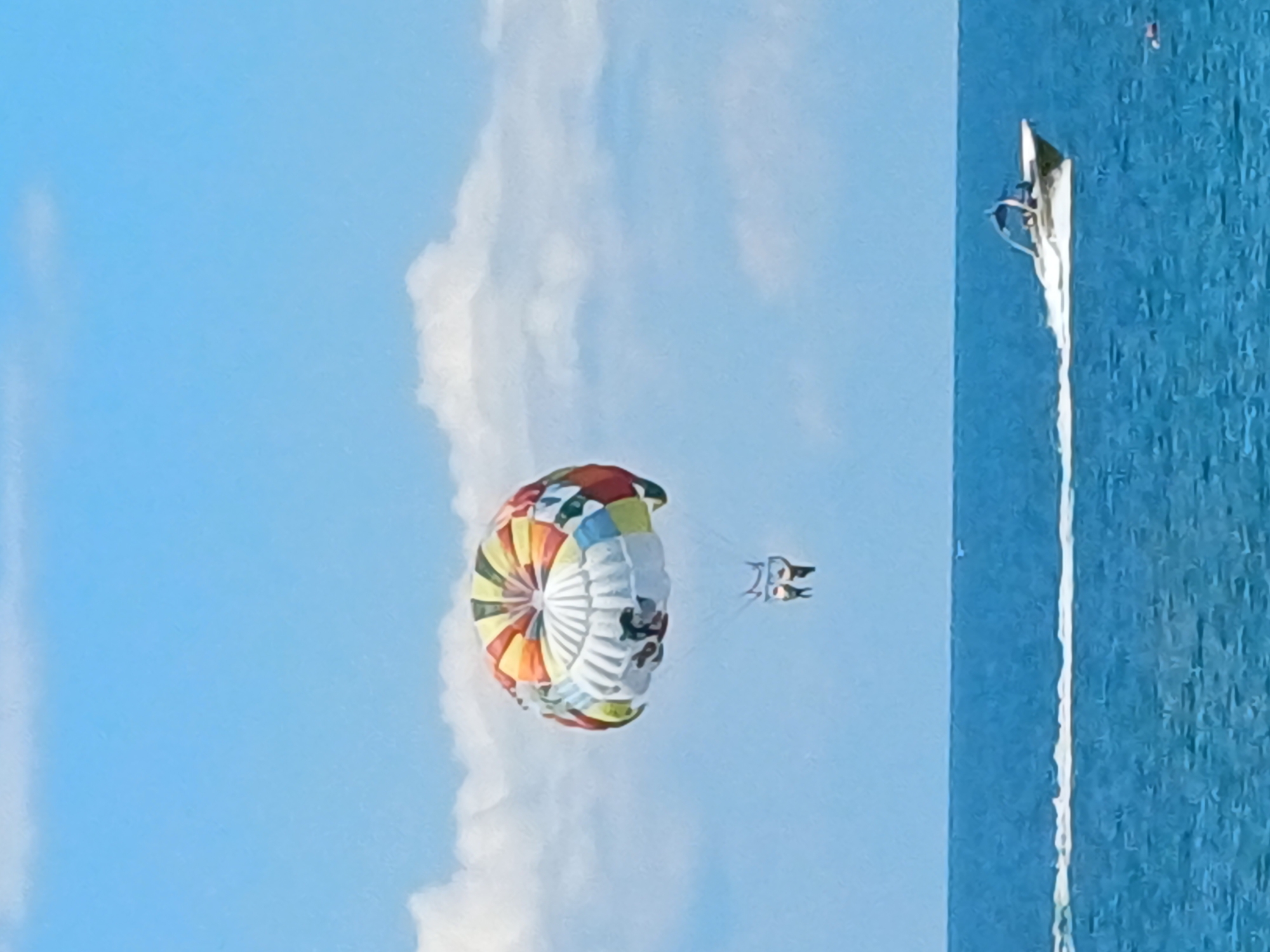 A parasailer is being towed by a boat in the azure waters of Sochi, Russia. The parasail is colorful with red, orange, yellow, green, blue, and white panels, and it has a white section with cartoonish figures depicted on it. Two people are seated in the harness below the parasail, appearing to be enjoying the ride. The boat, a sleek white speed boat, is actively moving through the water, creating a white wake behind it. The sea is a vibrant blue, and the sky above is a clear blue with scattered white clouds. The overall scene captures a recreational activity taking place on a sunny day.