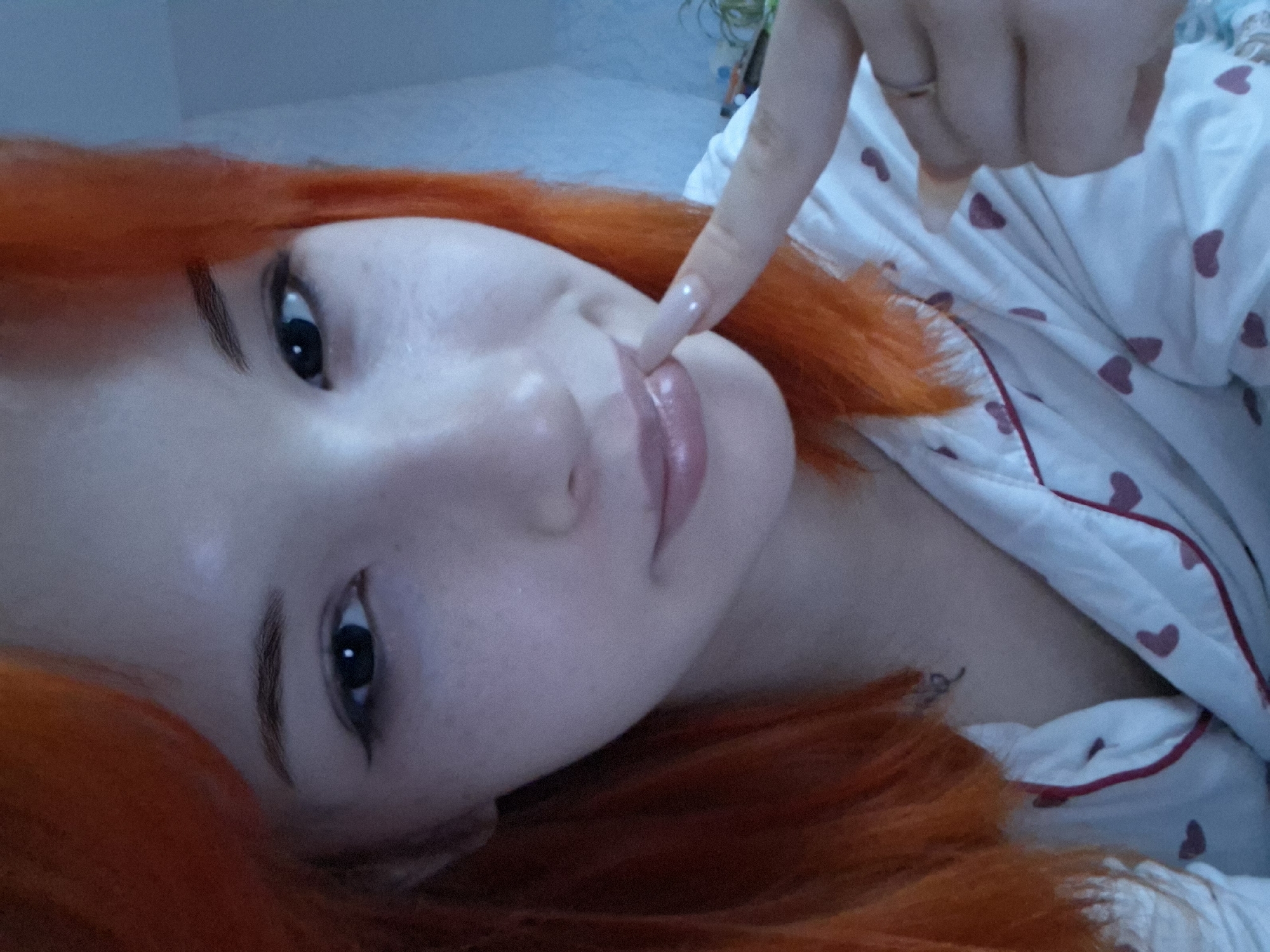 A person with red hair is lying down and touching their lip with a finger. The person is wearing white pajamas with red hearts. The setting appears to be indoors, possibly a bedroom. The location is unknown and not identifiable as Chaikovsky, Russia from the image.
