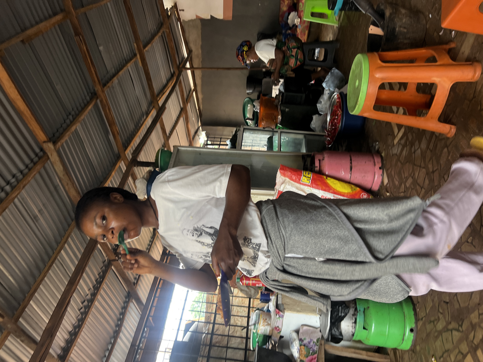 In a rustic, open-air structure in Nkpor, Nigeria, a young woman is brushing her teeth. She is wearing a white t-shirt with a graphic print and light purple pants, with a grey cardigan tied around her waist. In her right hand, she holds a green toothbrush, and in her left, a blue plate with food. The environment is cluttered, featuring various kitchenware, gas cylinders, and seating arrangements, indicating a functional, perhaps communal, living or working space. Sunlight filters through the corrugated iron roof, illuminating the scene. Another woman, wearing a colorful headscarf, is visible in the background, engaged in an activity at a counter. The floor is tiled with a mosaic pattern, and the overall impression is one of everyday life in a busy Nigerian locale.