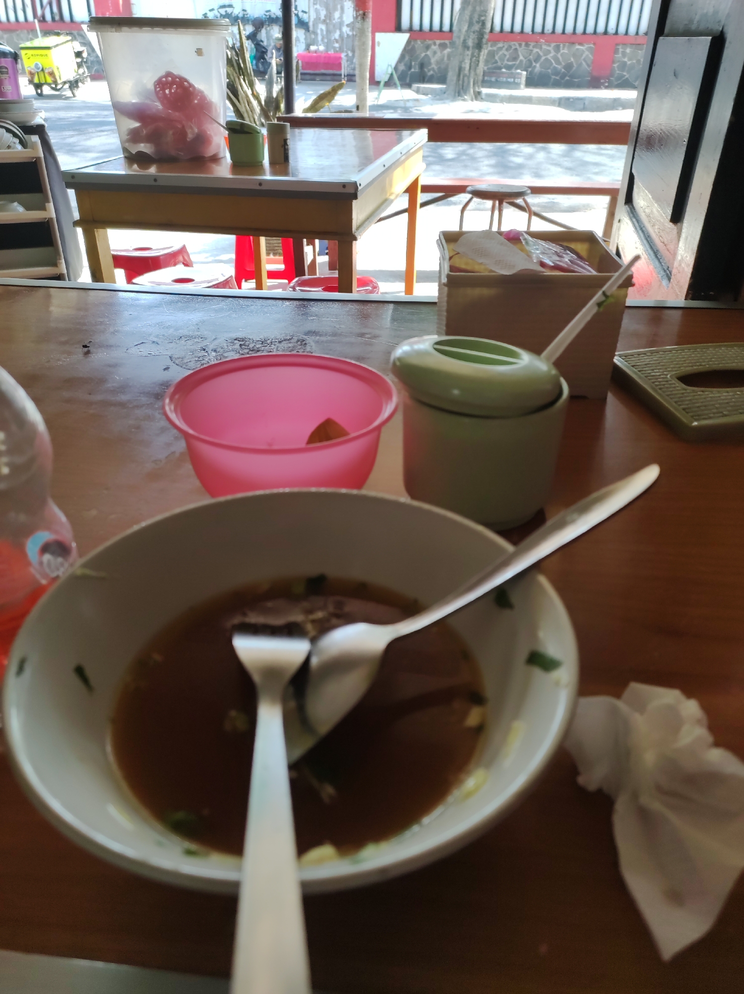 An overhead, close-up shot shows a wooden table with various items on it, suggesting an eating establishment in Blitar, Indonesia. In the foreground, a white bowl with a thick rim contains dark brown broth and some scattered green garnishes. A silver spoon and fork rest inside the bowl, partially submerged in the broth. To the left of the bowl, a clear plastic bottle with a red liquid is visible. Behind the bowl, a pink plastic bowl is positioned on the table, and next to it, a light green condiment dispenser with a lid. Further back, a beige rectangular box holds napkins and possibly other disposable items. The background reveals a window with a view of the outdoors, showing tables and chairs, a yellow food cart, and a building with a red and white facade. The overall impression is that of a casual dining setting.