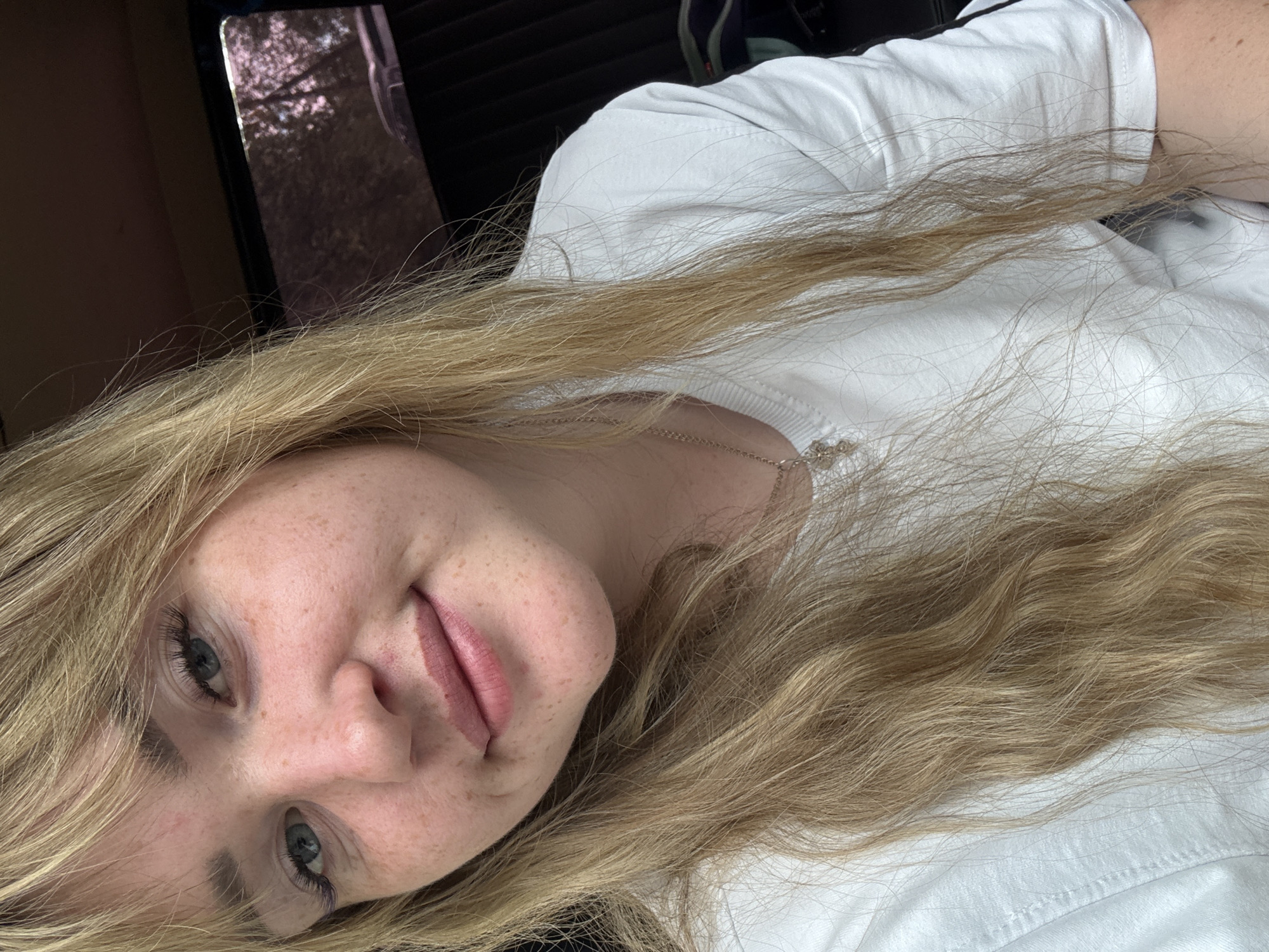 A young woman with blonde hair, blue eyes, and freckles is taking a selfie. She is wearing a white t-shirt and a necklace. She appears to be indoors, possibly in a car, as there is a window with trees visible in the background. The woman is smiling.