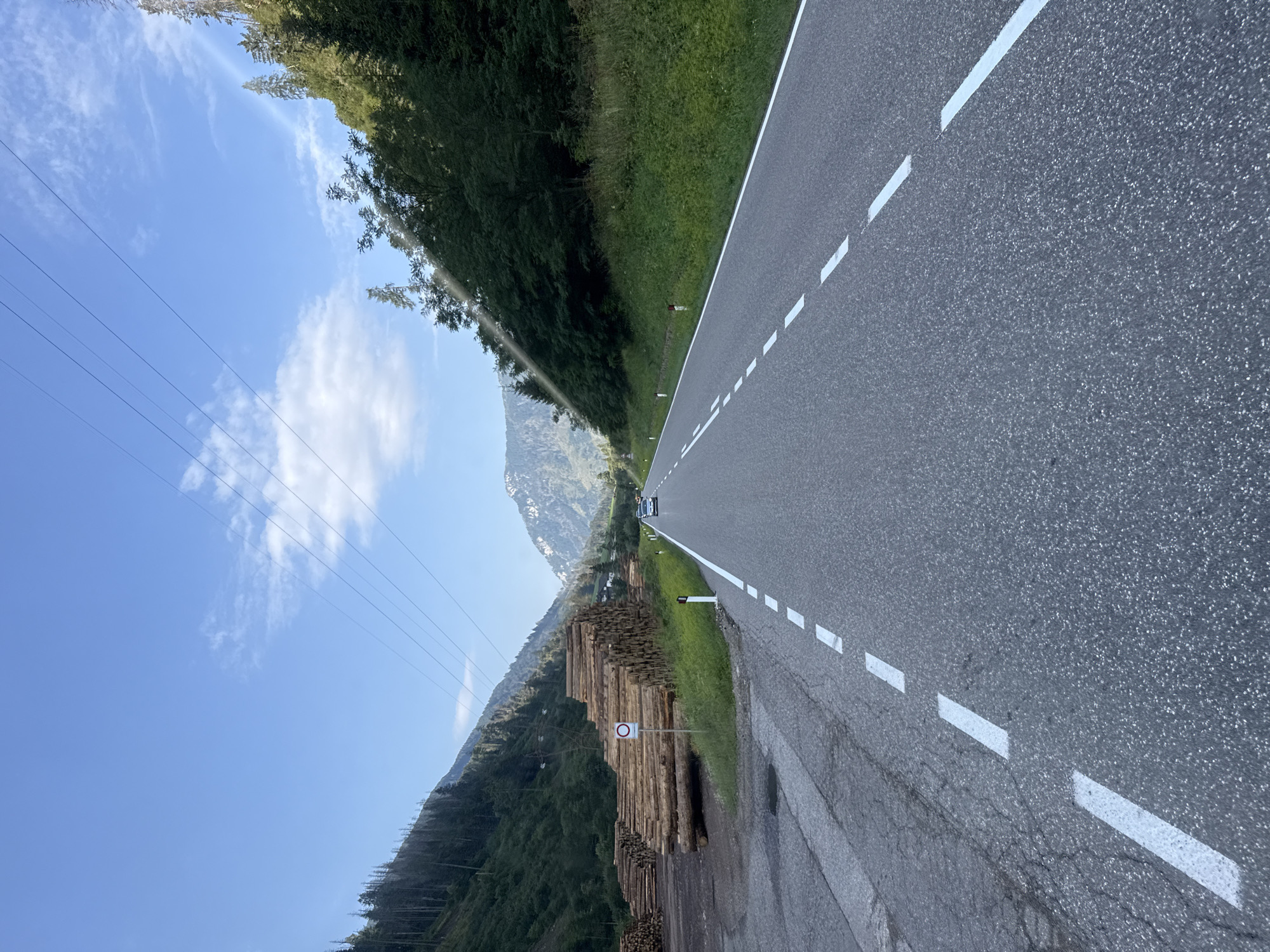The image shows a scenic mountain road in the vicinity of Moena, Italy. The road is paved and has white dashed lines indicating lanes. On the left side of the road, there is a large pile of stacked logs, suggesting a logging operation or wood storage area. A circular red and white sign with a diagonal bar is posted near the logs, likely indicating a restriction. Further up the mountain slopes, dense green forests cover the terrain. In the distance, majestic mountains with rocky peaks rise against a clear blue sky with scattered white clouds. Power lines are visible stretching across the sky. A light-colored car is driving away from the camera on the road. The environment suggests a rural, mountainous area known for its natural beauty and possibly forestry activities.
