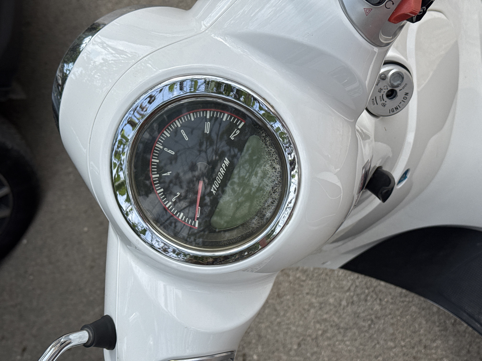 The image shows the dashboard of a white scooter. The speedometer reads around 2000 RPM. The ignition is nearby. The location (Budva, Montenegro) cannot be determined from the image.