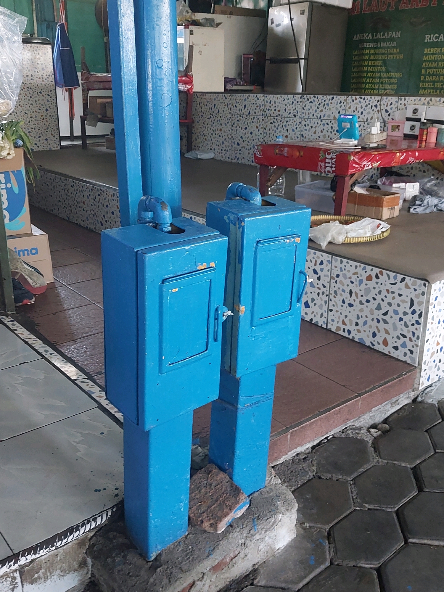The image captures an outdoor scene in what appears to be a food stall or restaurant area in Malang, Indonesia. In the foreground, two identical bright blue, rectangular metal boxes, possibly for trash or storage, stand side-by-side on a concrete foundation. Each box has a small hinged door with a simple latch. The boxes are mounted on thick, blue, square posts. The ground is a mixture of hexagonal dark gray paving stones and a concrete walkway.
In the middle ground, a red-covered table with a white and red patterned cloth is visible. On the table, a small blue electrical appliance, likely a mini iron, is plugged into a power strip. Several other items, including small white boxes, pink tissue boxes, and what looks like a bottle of water, are also on the table. Behind the table, a counter or wall is decorated with a mosaic of small, multi-colored tiles.
In the background, a large silver refrigerator is visible. Above the counter, a green sign with text in what appears to be Indonesian lists various food items. A blue cloth is hanging from a rack on the left side of the image, partially obscuring a chair. A stack of boxes is also on the left side of the image, with one box displaying the word "prima". The overall impression is a functional, somewhat utilitarian outdoor dining or service area.