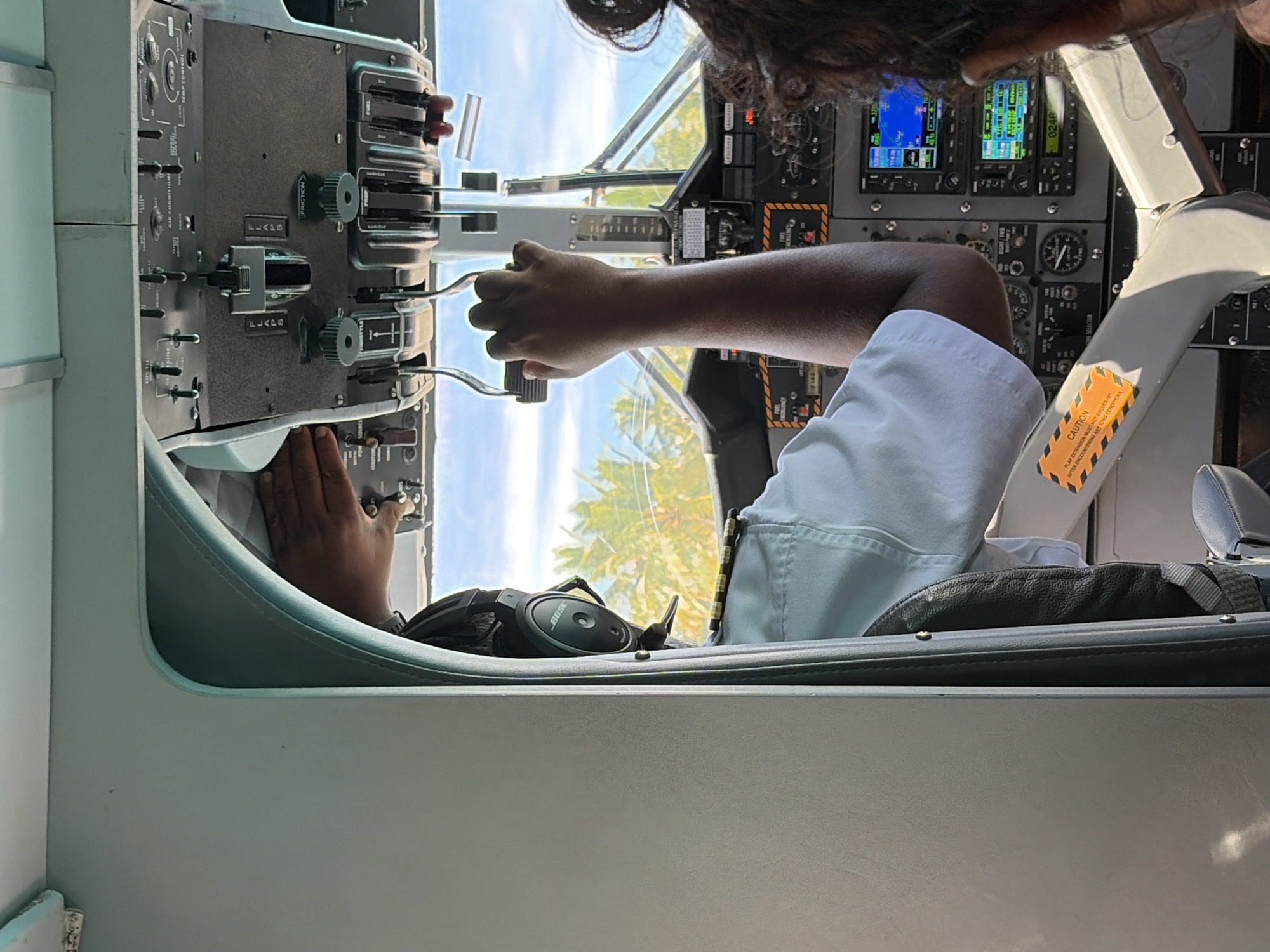 The image provides an interior view of an aircraft cockpit, likely taken from the passenger area, looking forward. A pilot, wearing a white uniform shirt with epaulets and a black headset with "BOSE" visible, is seated in the left seat, his right arm extended to operate the controls. His hand is gripping a lever, likely a throttle or power control. To the right of the pilot, a person's head and shoulder are visible in the foreground, their dark hair pulled back. The cockpit is filled with various instruments, gauges, and switches, indicative of a small aircraft. Prominently displayed on the panel are the "FLAPS" levers and engine control levers, labeled "THROTTLE," "PROP," and "FUEL." A bright orange "CAUTION" sticker is affixed to a part of the cockpit structure. Through the cockpit windows, a bright blue sky and lush green palm trees are visible, suggesting a tropical location. The presence of palm trees and the bright sky are consistent with the environment of Maalhos, Maldives, a known island destination in the Maldives archipelago. The lighting indicates it is daytime, with sunlight illuminating the scene. The overall impression is one of an active flight or pre-flight preparation.
