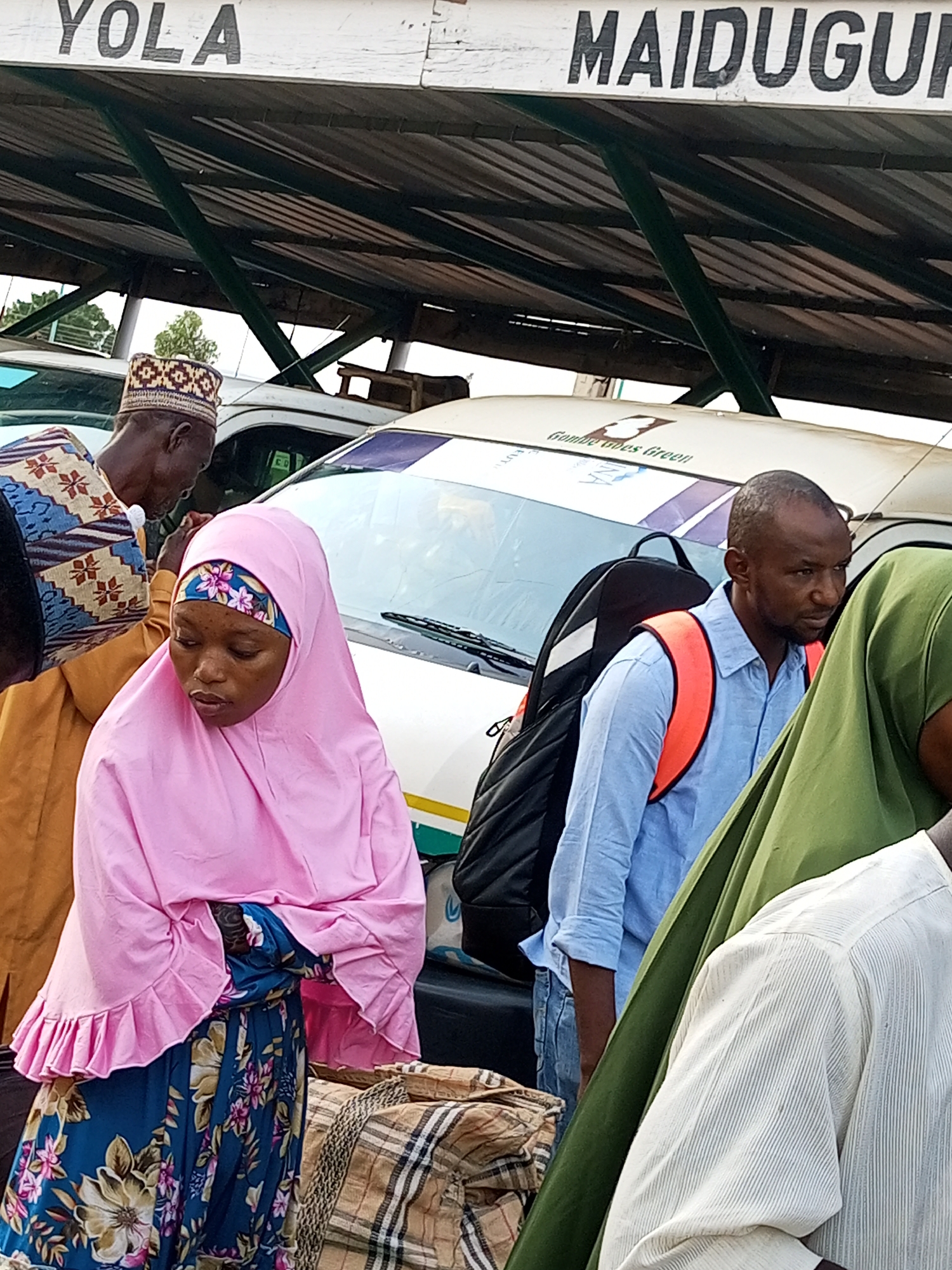 The image depicts a scene at a motor park, likely in Gombe, Nigeria, indicated by the signage "YOLA" and "MAIDUGURI" visible overhead, which are major cities in northeastern Nigeria. In the foreground, several people are gathered around vehicles. A woman wearing a pink hijab and a floral dress is looking downwards. Beside her, a man in a blue shirt with an orange reflective vest is looking to the side, carrying a black bag. Another person in a traditional brown and patterned outfit is partially visible on the left. The background shows the roof structure of the motor park and the windshields of parked vehicles. Bags and luggage are visible on the ground. The overall impression is of a busy transportation hub.