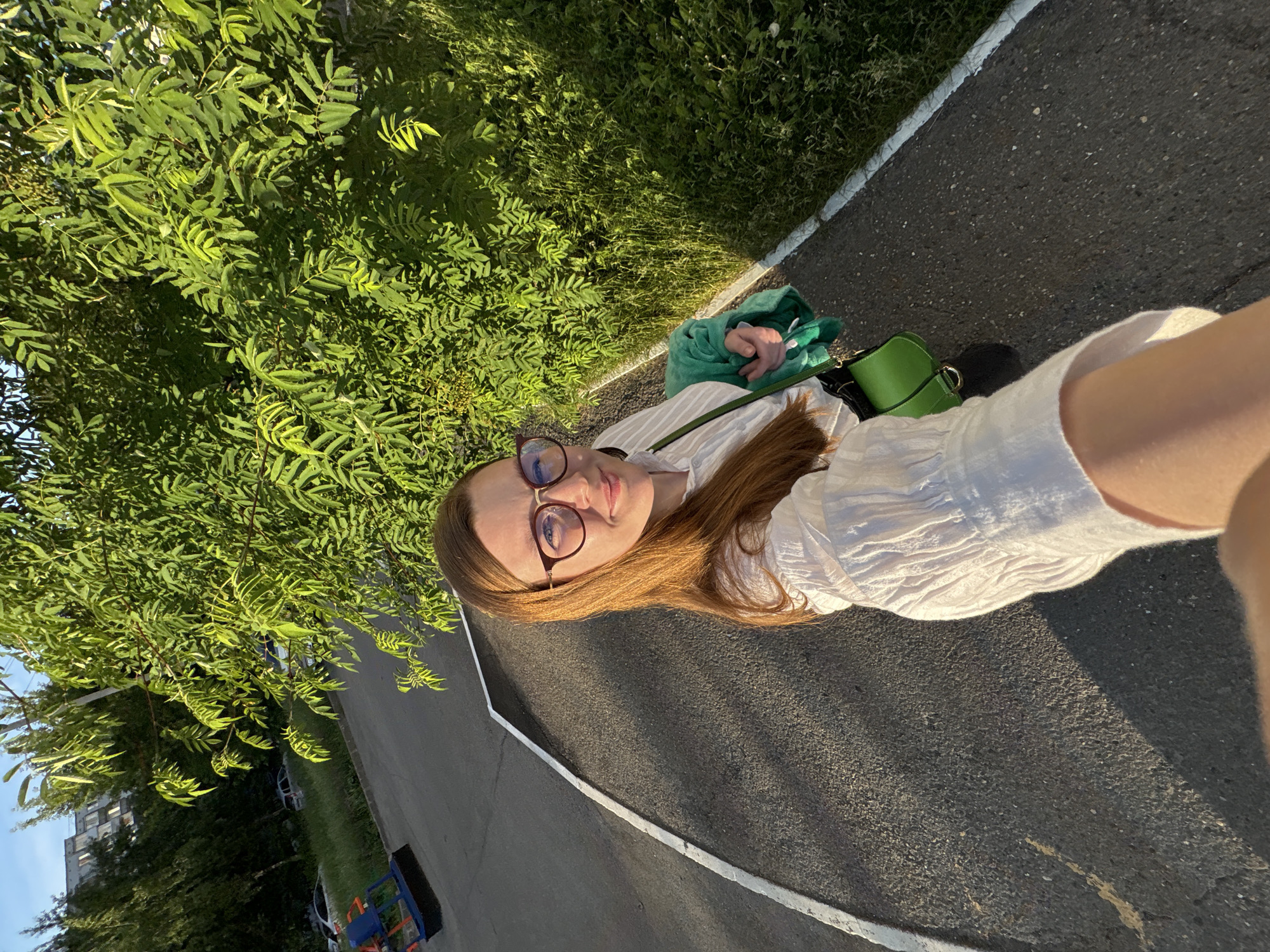 In Izhevsk, Russia, on a paved path bordered by white lines and a grassy area, a woman with long auburn hair and glasses is taking a selfie. She is wearing a white button-up shirt and carrying a green purse and a green blanket. Behind her, a lush green tree with vibrant leaves fills the frame. In the background, a building and parked cars are visible. The scene is bathed in the warm light of what appears to be late afternoon sun.
