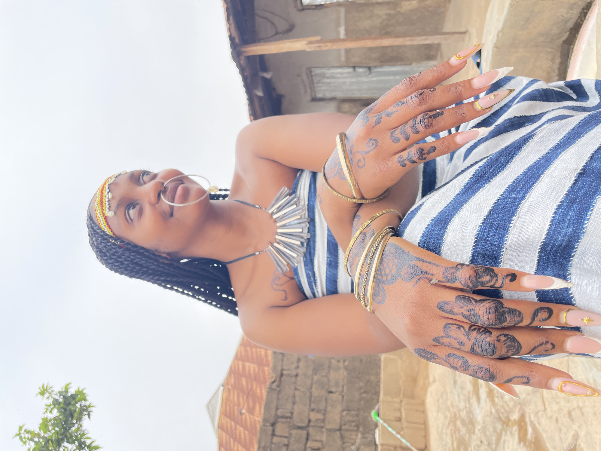 A person with braided hair and adorned with jewelry, including a nose ring, necklace, and several gold bracelets on each wrist, is photographed outdoors in what appears to be the city of Jos, Nigeria. The person's hands are decorated with dark henna-like patterns and elaborate, long, gold-accented nails. The individual is wearing a striped blue and white garment and is positioned against a backdrop of buildings with corrugated metal roofing and brick walls. The sky appears overcast, and a small tree is visible in the periphery.