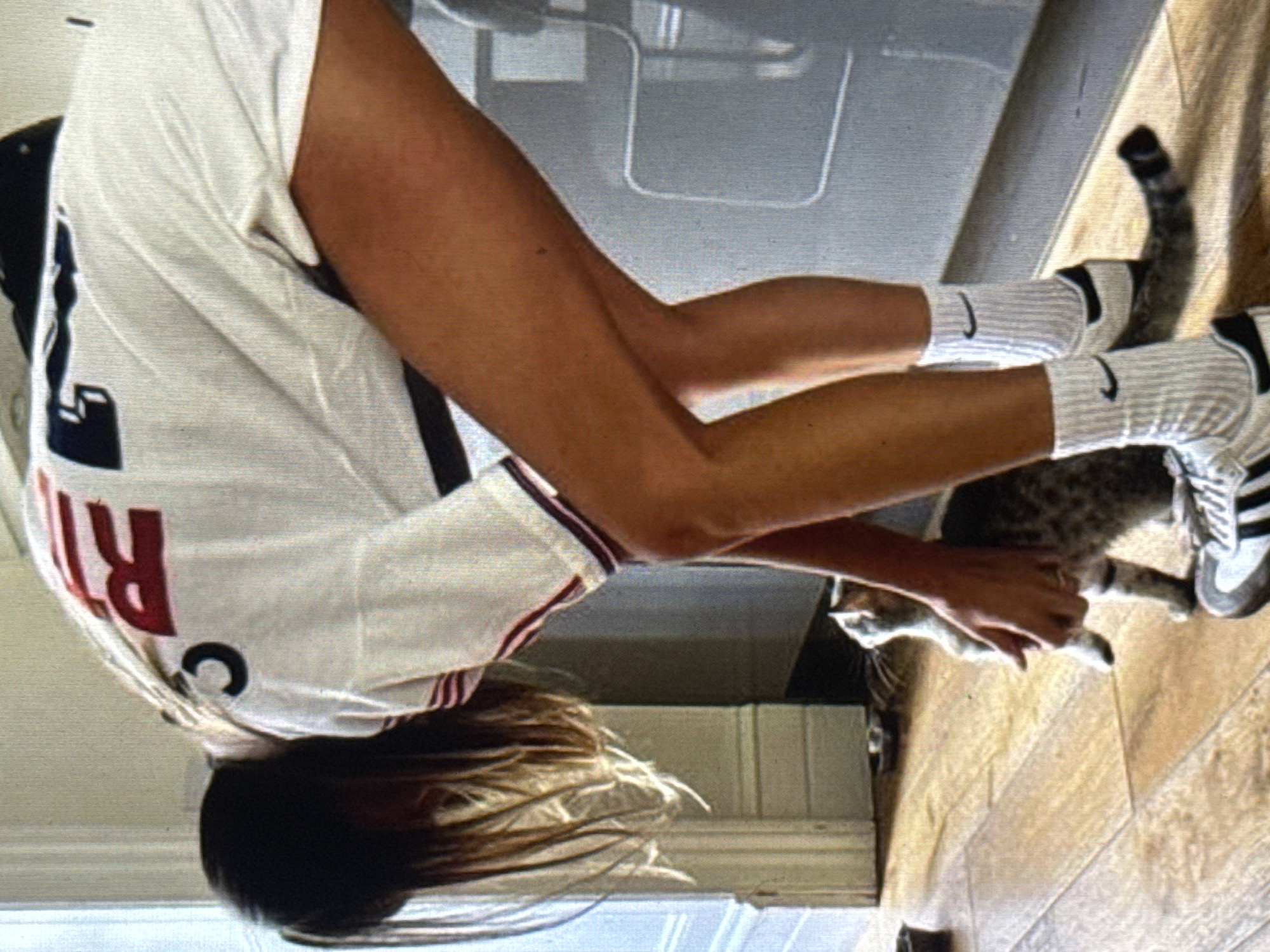 In Kuta Selatan, Indonesia, a person wearing a white t-shirt with red lettering and white socks with black stripes is interacting with a tabby cat on a wooden floor. The person is bending down, with their hands near the cat. The environment appears to be indoors, possibly a home, with a wall and furniture visible in the background. The lighting suggests it is daytime, with natural light filtering into the room. The scene is casual and domestic, with the person and cat engaged in a gentle interaction.