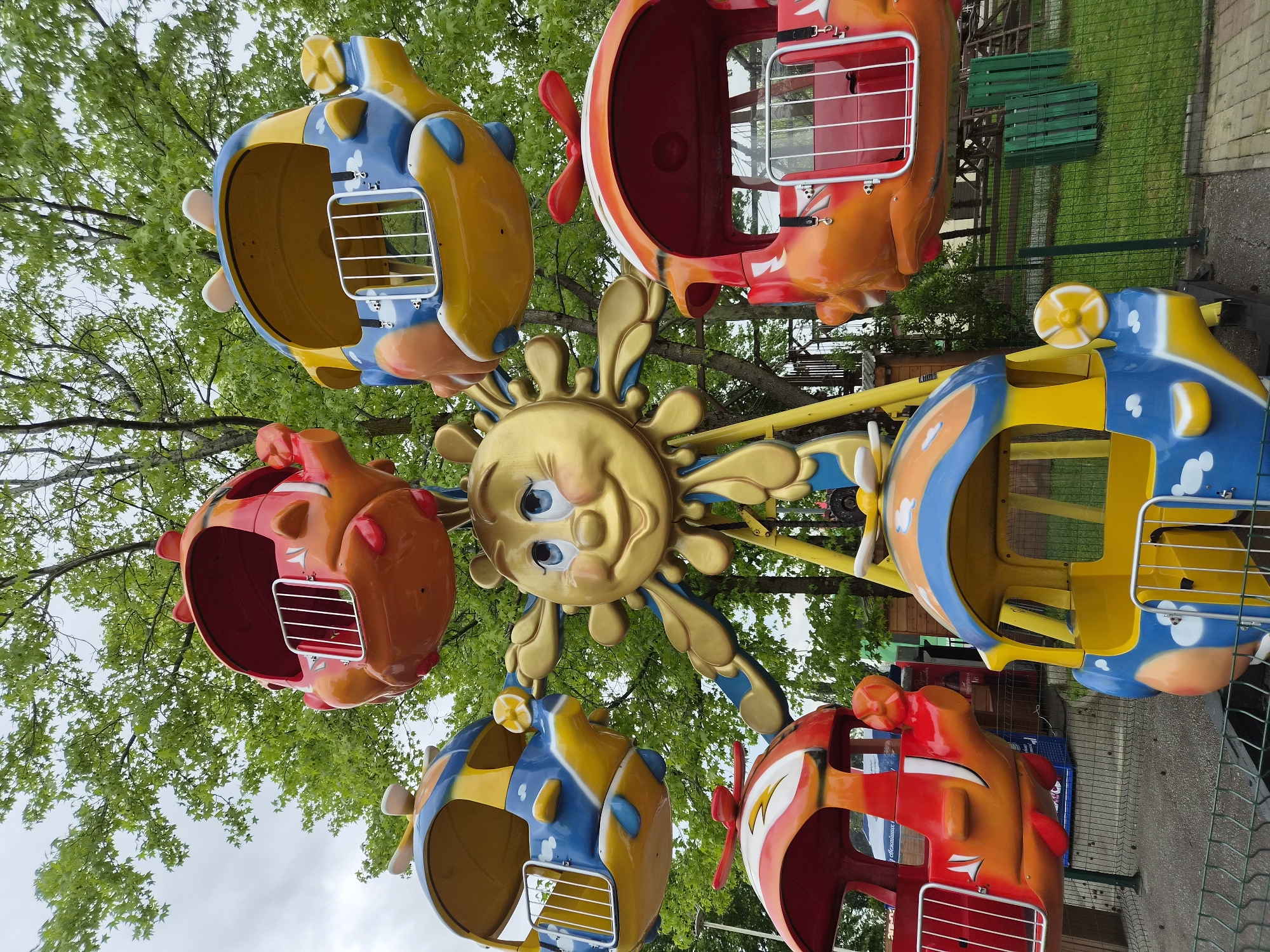 A colorful children's amusement ride is the focal point of the image. The ride features five airplane-shaped seats, each painted in different color combinations, including blue and yellow, orange and red, and red and white. The central part of the ride is shaped like a smiling golden sun. The ride appears to be set up in a park or recreational area, with green trees and a cloudy sky visible in the background. A green metal fence and benches are also visible. The location is in the city of Sochi, Russia.