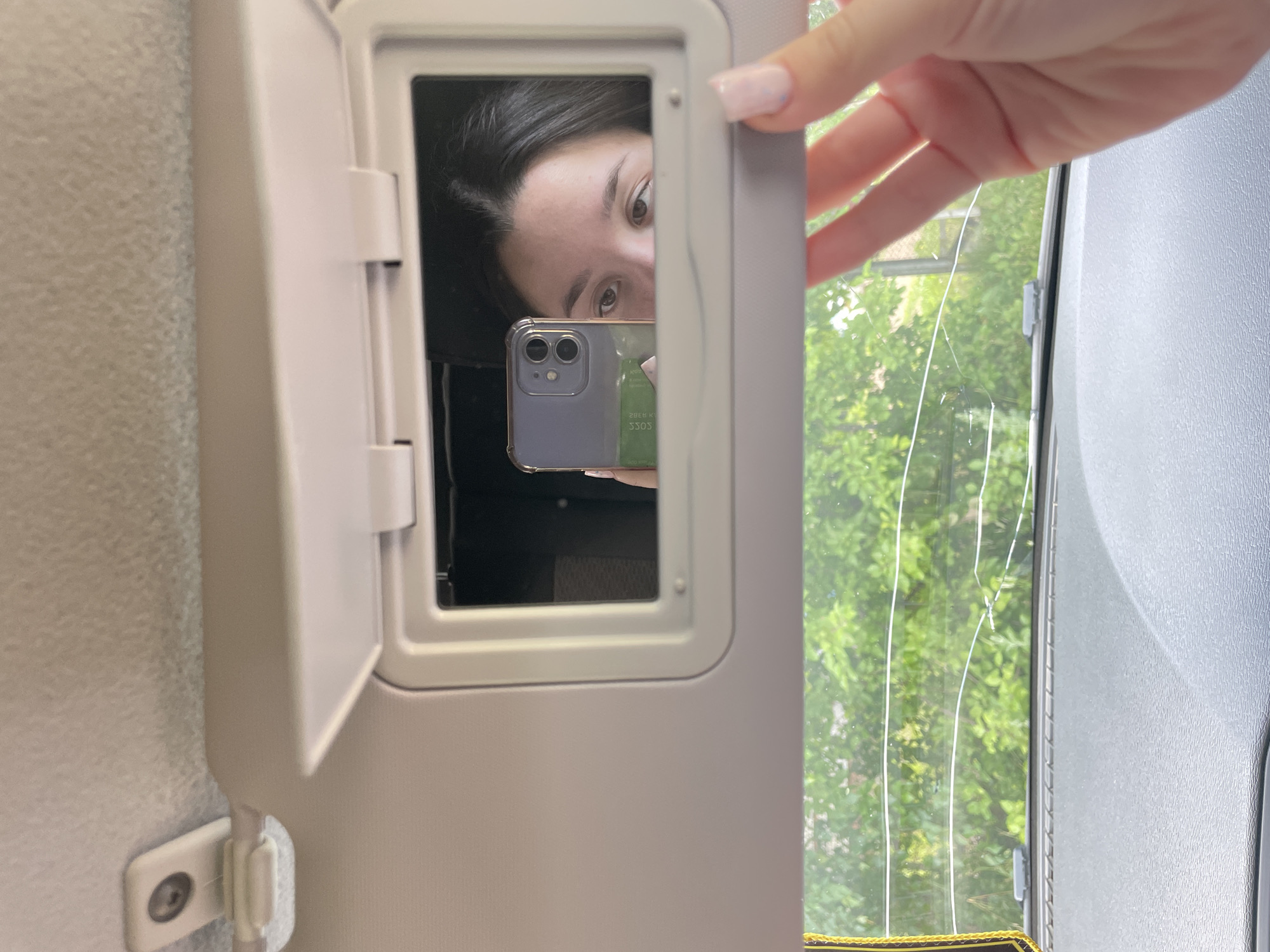 This media file shows a person's face reflected in a vanity mirror, which is part of a car's sun visor. The person, likely a young woman, is looking into the mirror with a neutral expression. She is holding a smartphone, the screen of which is also visible in the reflection, displaying what appears to be a green card with text. A hand with painted nails is visible in the upper right corner of the frame, reaching towards the edge of the sun visor. The sun visor is open, revealing the mirror and a glimpse of the car's interior. Outside the car, through a window with visible cracks, lush green trees and foliage are apparent, suggesting an outdoor setting during daylight hours. The overall lighting indicates it is daytime, possibly mid-morning or afternoon, with bright sunlight. The scene appears to be inside a vehicle. No specific action or event is depicted other than the act of looking in the mirror and the hand reaching out. The visible text on the phone screen is upside down and reversed, reading "3303 HORE" or similar. The location is noted as Nozhovka, Russia.