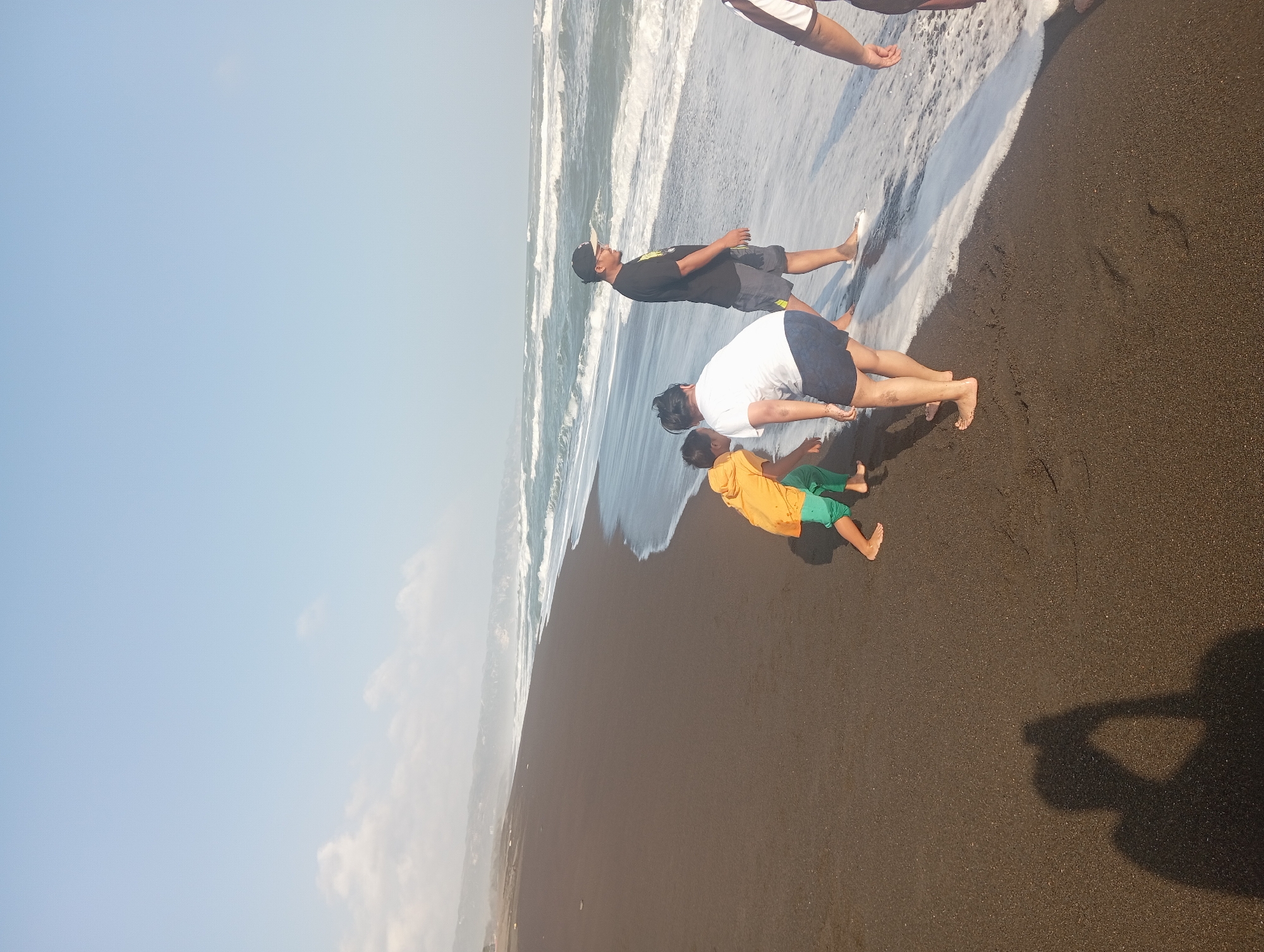 The image captures a sunny day at Parangtritis beach in Indonesia, with a dark sand beach stretching towards the ocean. Four people are present, with three in the foreground and one more partially visible on the right. In the center, a person wearing a white t-shirt and dark shorts is bending down, facing away from the camera, towards two children. One child, wearing an orange hooded jacket and green shorts, is also bending down. The other child, wearing a white t-shirt and blue shorts, is standing. To the right of them, a man in a black t-shirt and dark shorts stands, looking towards the water. He is wearing a black cap. The ocean has moderate waves and white foam washing onto the shore. In the distance, the coastline is visible with a hazy, hilly landscape. The sky is clear and blue with a few scattered clouds. The shadow of the person taking the photograph is visible on the sand in the lower left corner.