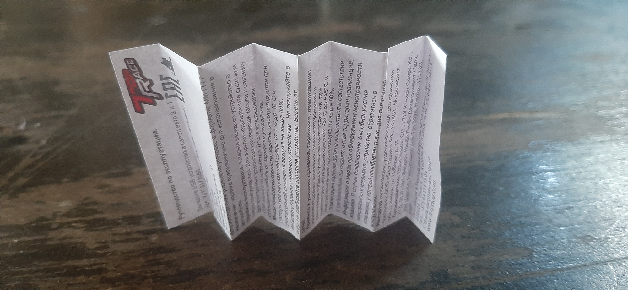 This is a close-up, eye-level shot of a folded piece of paper, likely an instruction manual or warranty information, lying on a rustic wooden surface. The paper is white and appears to be printed with black and red text, as well as a logo that reads "TRACE." The paper is creased in a zig-zag pattern, suggesting it's a multi-panel leaflet. The wooden surface has a visible grain and appears to be aged, with some light and shadow play suggesting diffused lighting, possibly from an indoor setting during the day.
The text on the paper is in Russian and appears to be technical information and contact details. It includes phrases like "Руководство по эксплуатации" (Instruction manual), "Зарядное USB устройство" (USB charging device), and details about its use, storage, and manufacturer. The logo "TRACE" is prominent on the left side of the visible paper. The overall impression is of a functional document presented in an informal setting. There are no people visible in the image.