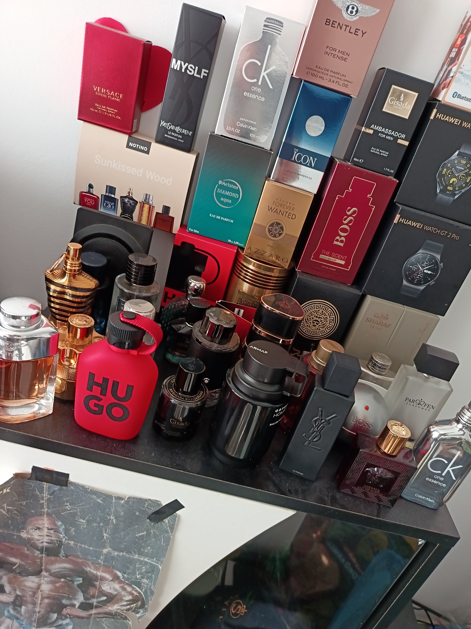 A high-angle, medium shot captures a collection of assorted perfume bottles and their boxes arranged on a dark shelf. The items are predominantly masculine fragrances, featuring brands such as Versace, Bentley, CK One, Boss, and Hugo. Some boxes are stacked, while others are placed side-by-side. The bottles vary in size, shape, and color, with prominent gold, red, and black hues.
In the background, a large, dark speaker is partially visible, with a printed poster of a muscular man taped to a white surface above it. The poster is partially obscured by the perfume collection and appears to be aged. The overall setting suggests a retail display or a personal collection. The lighting is somewhat dim, creating shadows that add depth to the arrangement.