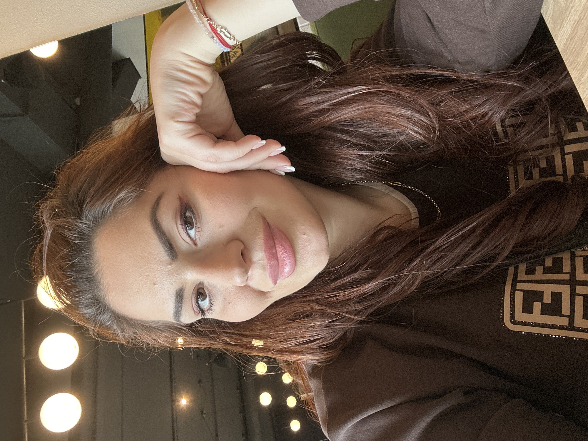 A selfie photograph of a woman indoors is presented. The woman has dark brown hair and fair skin. She is wearing a brown top with a logo pattern on the right side. Her left hand is raised to her face, with manicured nails. A beaded bracelet adorns her wrist. The background features multiple light bulbs hanging from the ceiling, suggesting the setting might be a cafe or restaurant. Based on the provided context, the location is Wrocław, Poland.