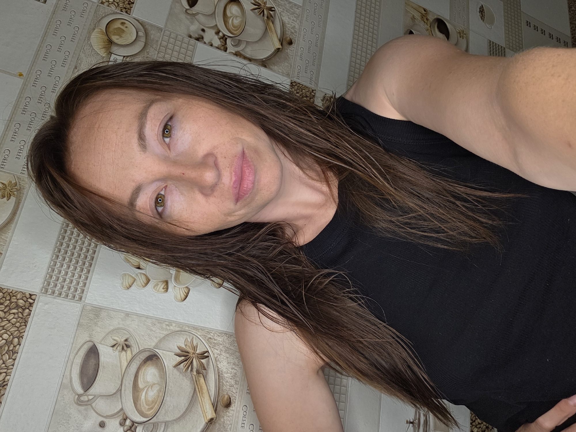 A woman with long brown hair, wearing a black tank top, looks directly at the camera. Her skin has freckles, and she has light brown eyes and pink lips. She is positioned in front of a wall covered in a tiled pattern featuring images of coffee cups, beans, and the word "COFFEE" repeated in a pattern. The lighting suggests an indoor setting, likely a kitchen or dining area. There is no indication of the specific location within Chaikovsky, Russia, or any specific action or interaction taking place beyond the portrait itself.