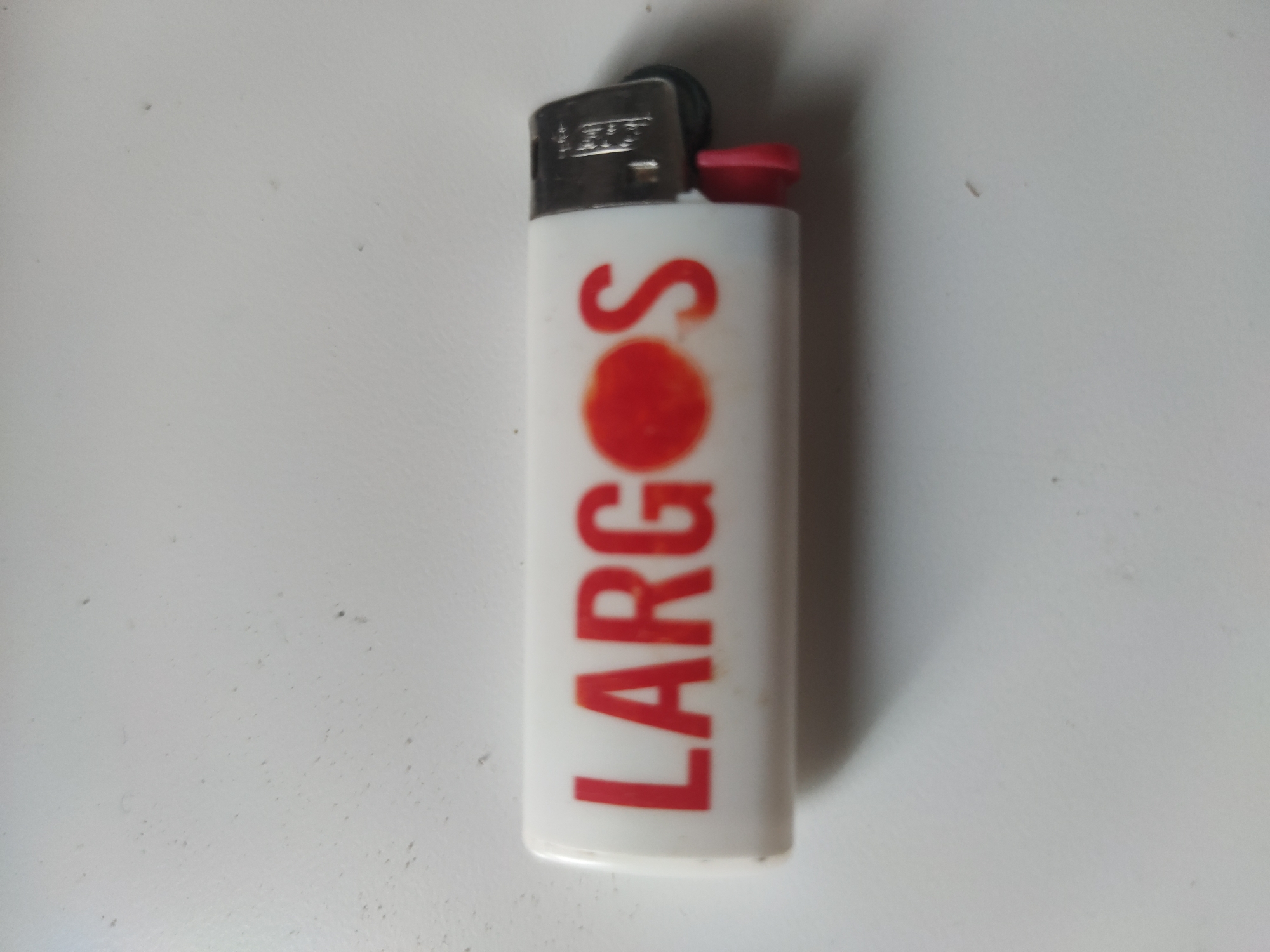 A white disposable lighter with the word "LARGOS" printed in red, vertically, is positioned on a white surface. The "O" in "LARGOS" is a red circle. The lighter has a silver-colored metal top and a red button. The environment is a plain, light-colored surface with some minor discoloration. The location is not identified.