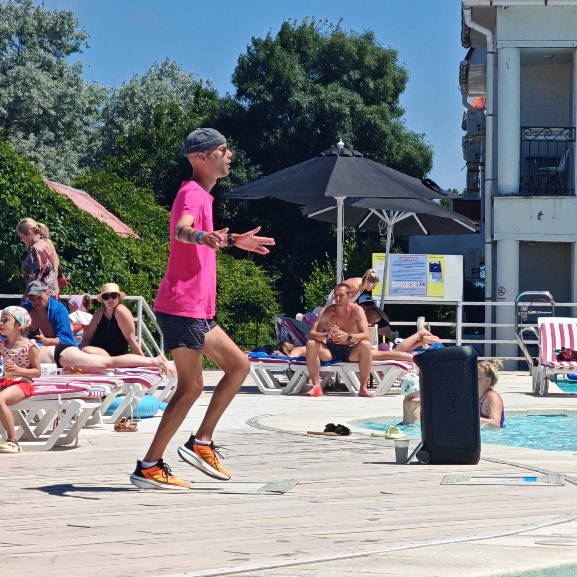 An outdoor daytime scene shows a group of people by a swimming pool. In the foreground, a person in a bright pink t-shirt and dark shorts with white stripes is jogging on a wooden deck. Their arms are outstretched as if dancing or exercising. A large black speaker is positioned near the edge of the pool to the right of the jogging person. Several people are lounging on deck chairs. One person in the background is partially visible in the pool. Trees and a building with balconies are visible in the background. The sky is clear and blue.