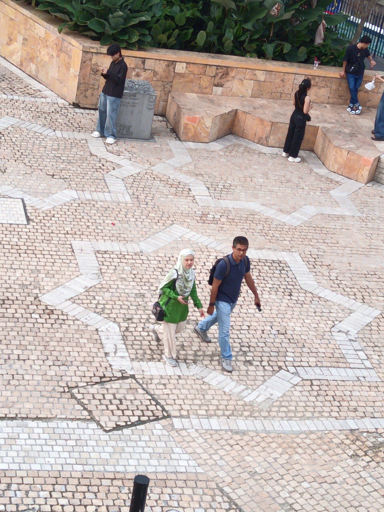 This aerial image captures an outdoor scene in Kuala Lumpur, Malaysia, likely in an urban plaza or park area. The ground is paved with light-colored bricks arranged in a patterned mosaic, featuring geometric white accents. In the background, a low stone wall with a textured, sandy-colored finish is visible, topped with lush green foliage.
Several individuals are present:
In the foreground, a couple is walking. The man, wearing a dark blue t-shirt, light blue jeans, glasses, and a backpack, walks with his right arm slightly extended, holding a dark object, possibly a phone. The woman beside him wears a green hijab, a green top, and light-colored pants. She carries a dark shoulder bag.
Further back, a man in a black long-sleeved shirt and blue jeans stands leaning against a grey concrete structure. He appears to be holding a cigarette. He is wearing white shoes.
To his right, a woman in black pants and a black top, with her hair pulled back, is also present, facing away from the camera. She is wearing white shoes and carrying a dark bag.
Another man in a black t-shirt and blue jeans stands near the woman in black, leaning on the stone wall. He is also wearing white shoes and has a dark bag slung across his body.
A part of a black pole is visible in the lower right corner of the frame. The overall impression is of a public space with people going about their day.