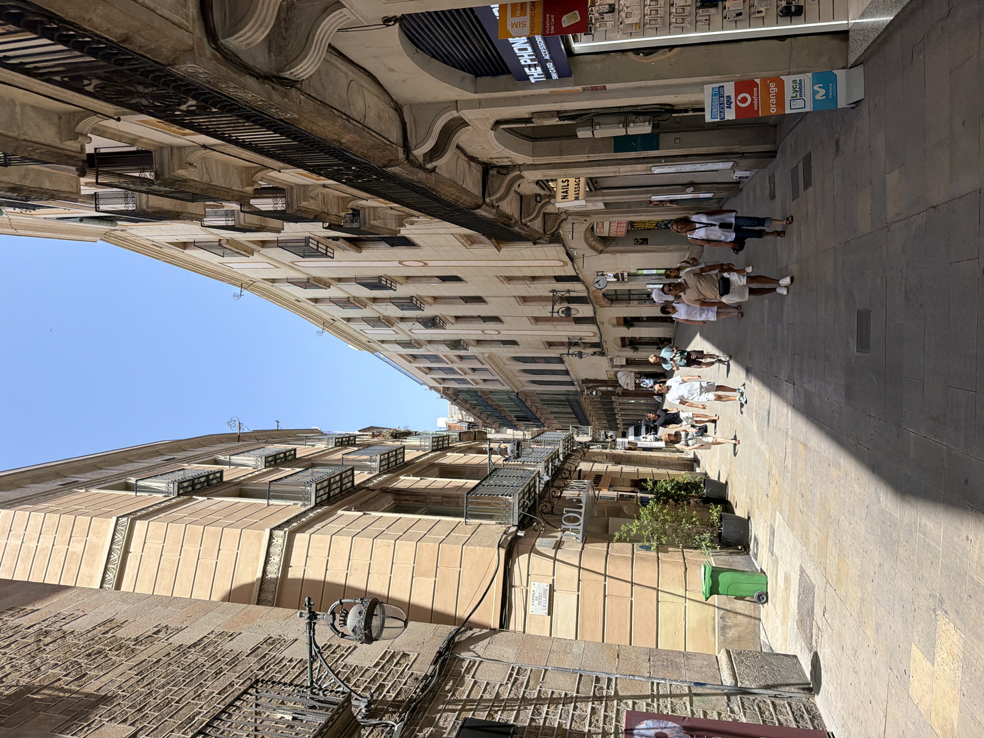 The image shows a narrow street in Barcelona, Spain, lined with tall, multi-story buildings. The buildings on both sides feature traditional architecture with balconies, ornate detailing, and light-colored facades. People are walking along the paved sidewalk, which is illuminated by sunlight casting shadows. Several shops and businesses are visible at street level, including one with signage for "THE PHONE" offering SIM cards and accessories, and another with "NAILS MASSAGE" advertisements. A green trash bin is located on the left side of the street near the corner of a building. The sky is clear and blue.