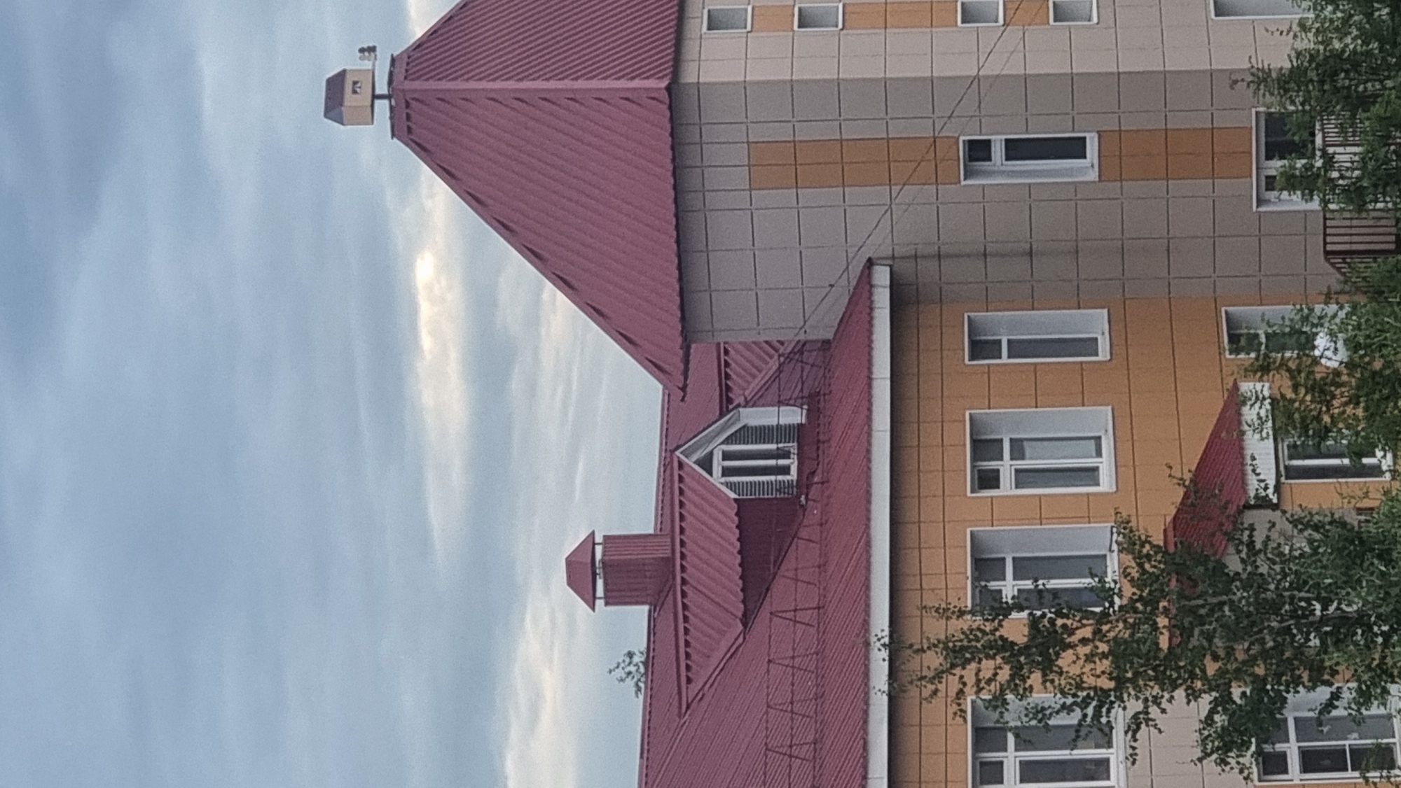This is a medium-shot, eye-level, outdoor photograph taken from an elevated perspective, looking at the upper floors and rooftops of buildings under a cloudy sky. The foreground features a large building with a red, corrugated metal roof. A prominent tower with a red, hip roof is attached to the main building. The tower's facade is clad in a grid of beige and light tan square tiles, with a vertical band of tan tiles running up the center. Several rectangular windows with white frames are visible on the tower's facade, some are dark, others are lighter suggesting interior lighting or reflections.
To the left of the tower, another section of the building has a similar red roof. Dormer windows with white trim and dark panes are visible on this section. The walls of this part of the building are clad in textured, yellowish-orange panels. More white-framed windows are visible on this facade.
At the very peak of the tower's roof, a small, house-like structure with a dark roof and light-colored walls sits atop a pole. A dark, weather vane with a shape resembling a person is attached to the side of this structure.
Green trees with dense foliage are partially visible in the lower left and right corners of the image, obscuring some of the lower parts of the buildings. The sky is overcast with visible layers of clouds, some of which are darker and appear heavy, while others are lighter and allow some ambient light to filter through. The overall scene suggests an urban or suburban environment.