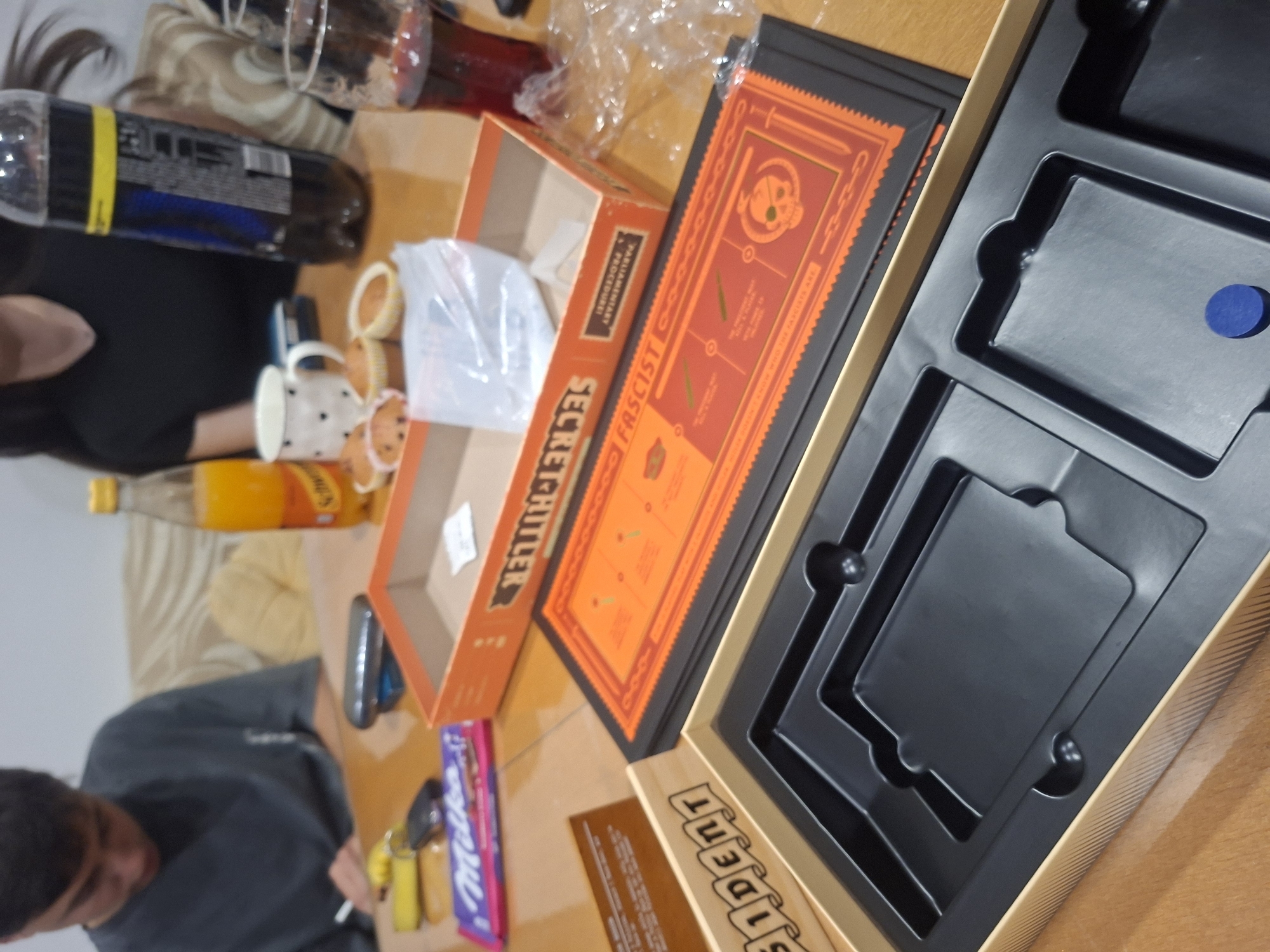 The image shows a table with a board game called "Secret Hitler", its box, and game components. Several people are seated at the table, seemingly in the middle of a game. There are also drinks, snacks, and personal items scattered around the table. The location, Gabrovo, Bulgaria, is not discernible from the visual information in the image.