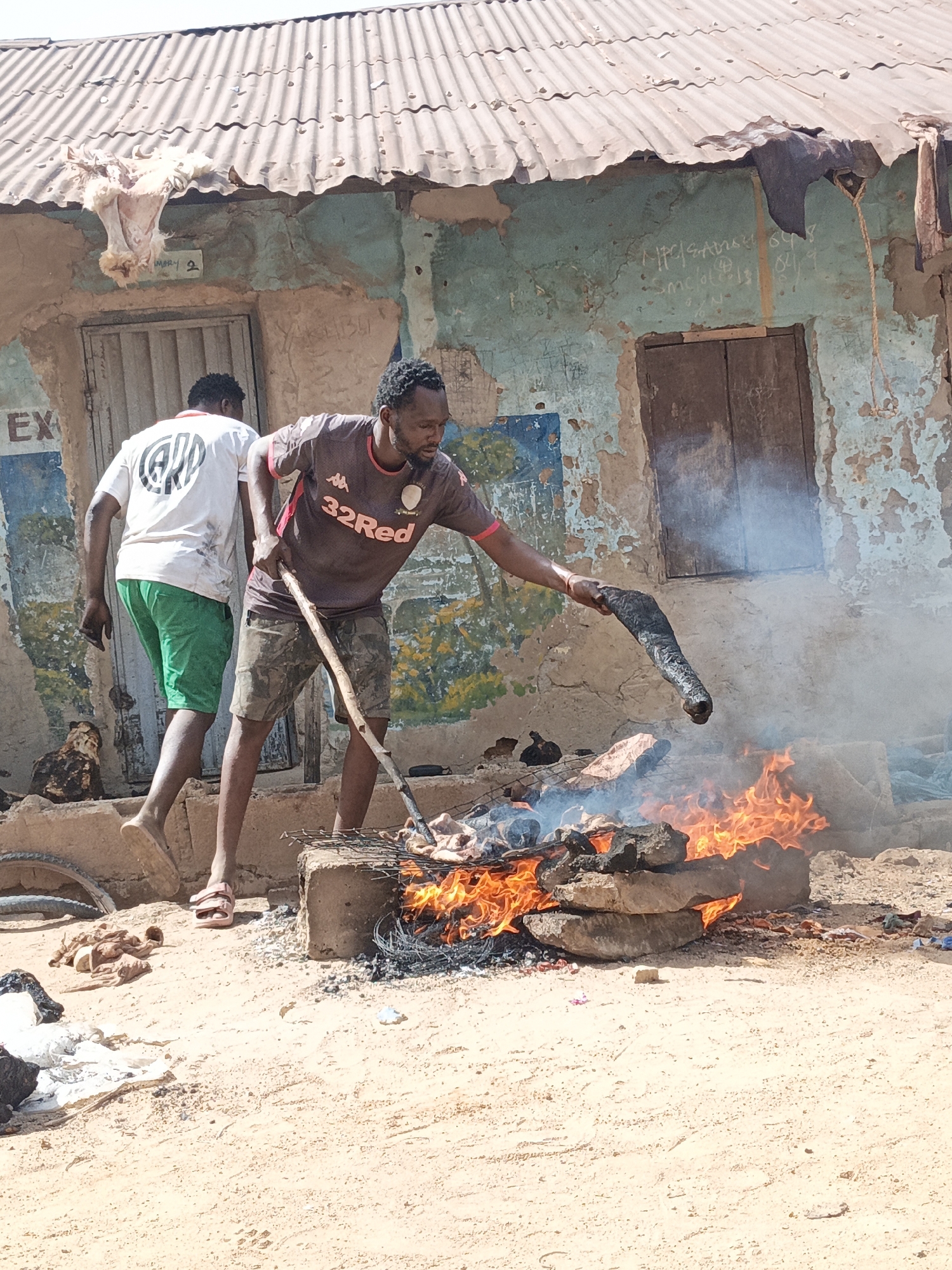 In Gwagwarwa, Nigeria, two men are outside a weathered building with a corrugated iron roof. One man, wearing a maroon and brown soccer jersey and camouflage shorts, stands by a fire constructed of stacked stones and cinder blocks, using a long stick to tend to something on a grill over the flames. He is holding a charred object aloft. The other man, in a white shirt and green shorts, stands to the side, seemingly observing. On the building, there is peeling paint, exposed brick, a boarded-up window, and graffiti. Parts of a hide hangs from the building. A bicycle wheel, debris, and a few domestic fowl are scattered around the sandy ground.