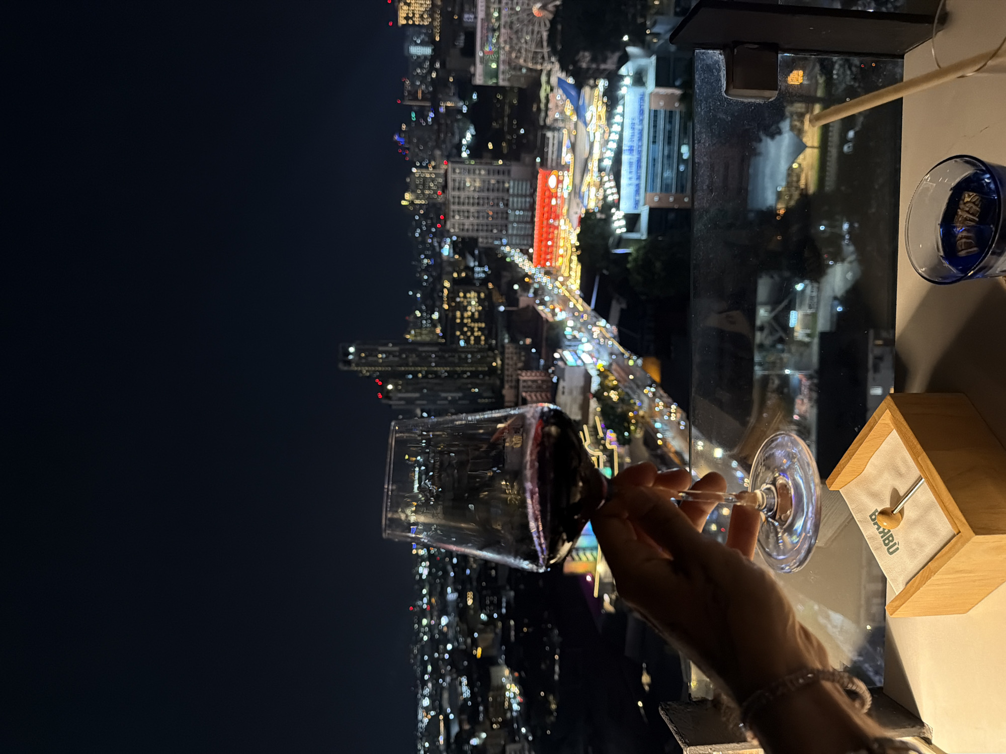 A nighttime view from a high vantage point shows a cityscape. A wine glass containing red wine is held in a hand, obscuring some of the buildings below. The city has many illuminated buildings and roads. To the right of the wine glass, a small, wooden box with a white cloth insert is visible. A drink in a blue-tinted glass stands near the box. The scene is set at a high altitude, likely a balcony or a window in Pattaya City, Thailand, given the combination of urban development and lit cityscapes.